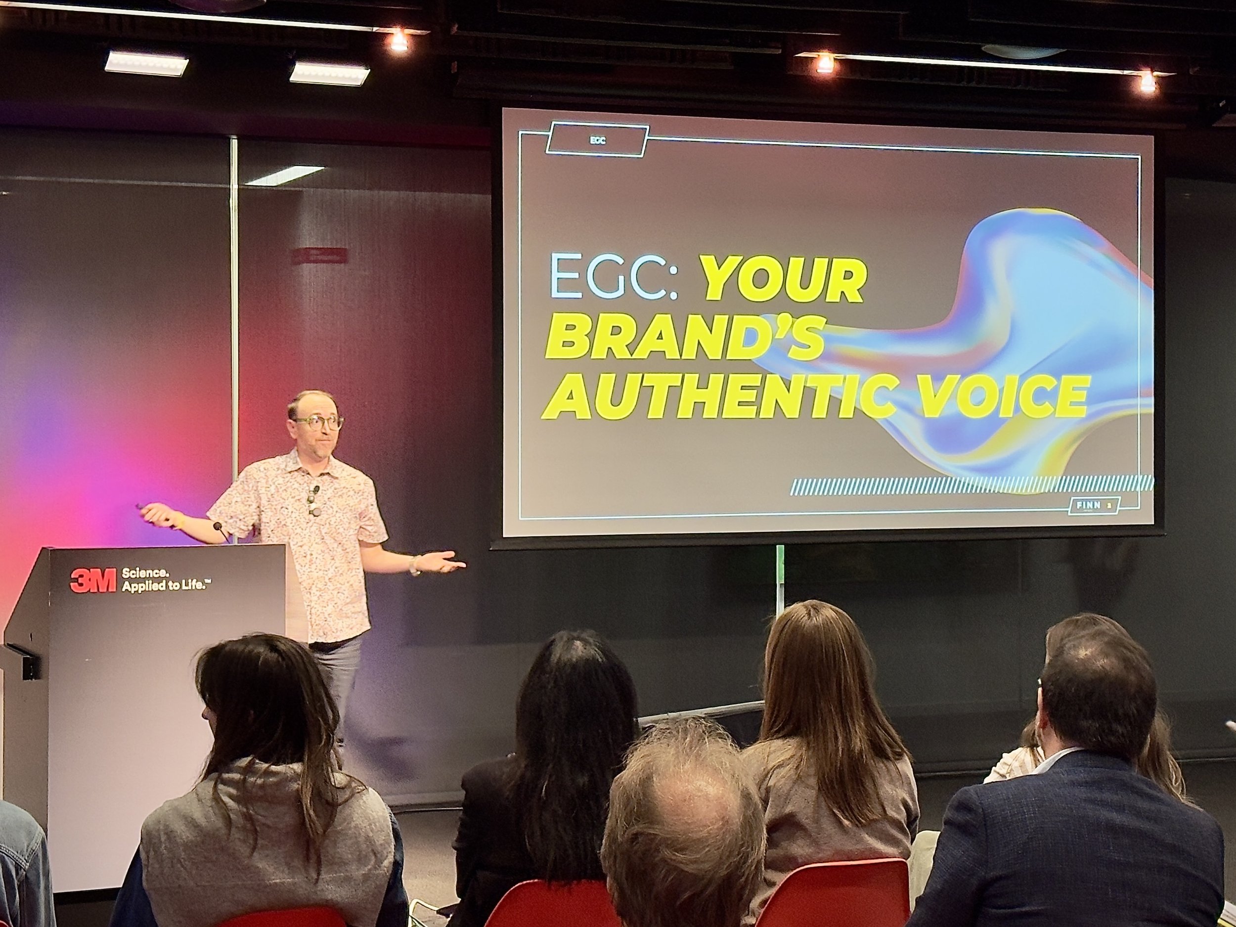 A man giving a presentation in a conference room with a large screen displaying the title 'EGC: YOUR BRAND'S AUTHENTIC VOICE'. The audience is seated facing the presenter and the screen, with a 3M logo on the lectern.