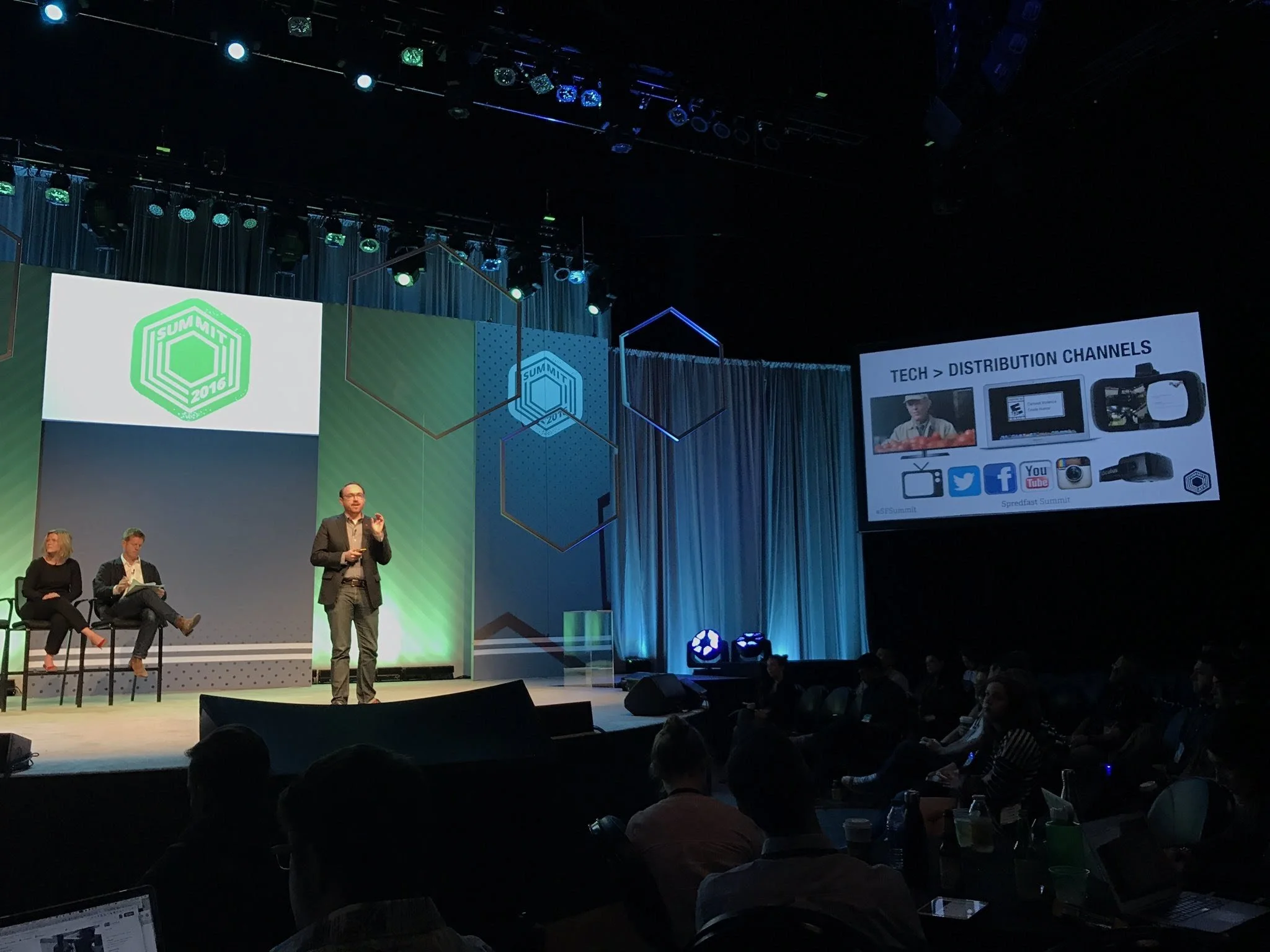 A conference or presentation at the 2016 Spreadfast Summit, with a speaker on stage, additional panel members seated to the left, and a large screen displaying a slide on distribution channels including social media icons.