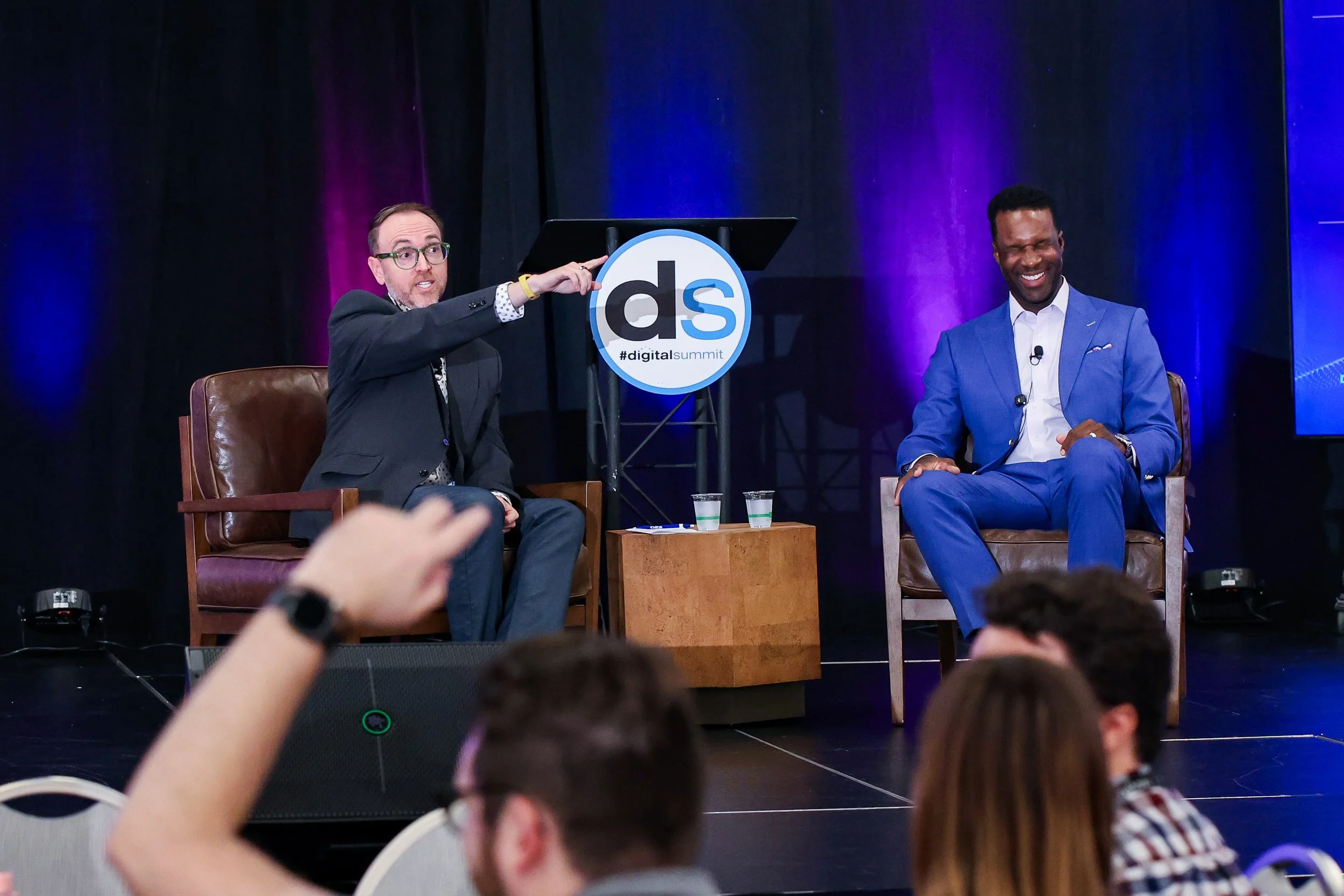 Two men seated on stage during a panel discussion at a digital summit, with one man speaking and the other smiling, in front of a backdrop with purple and blue lighting.