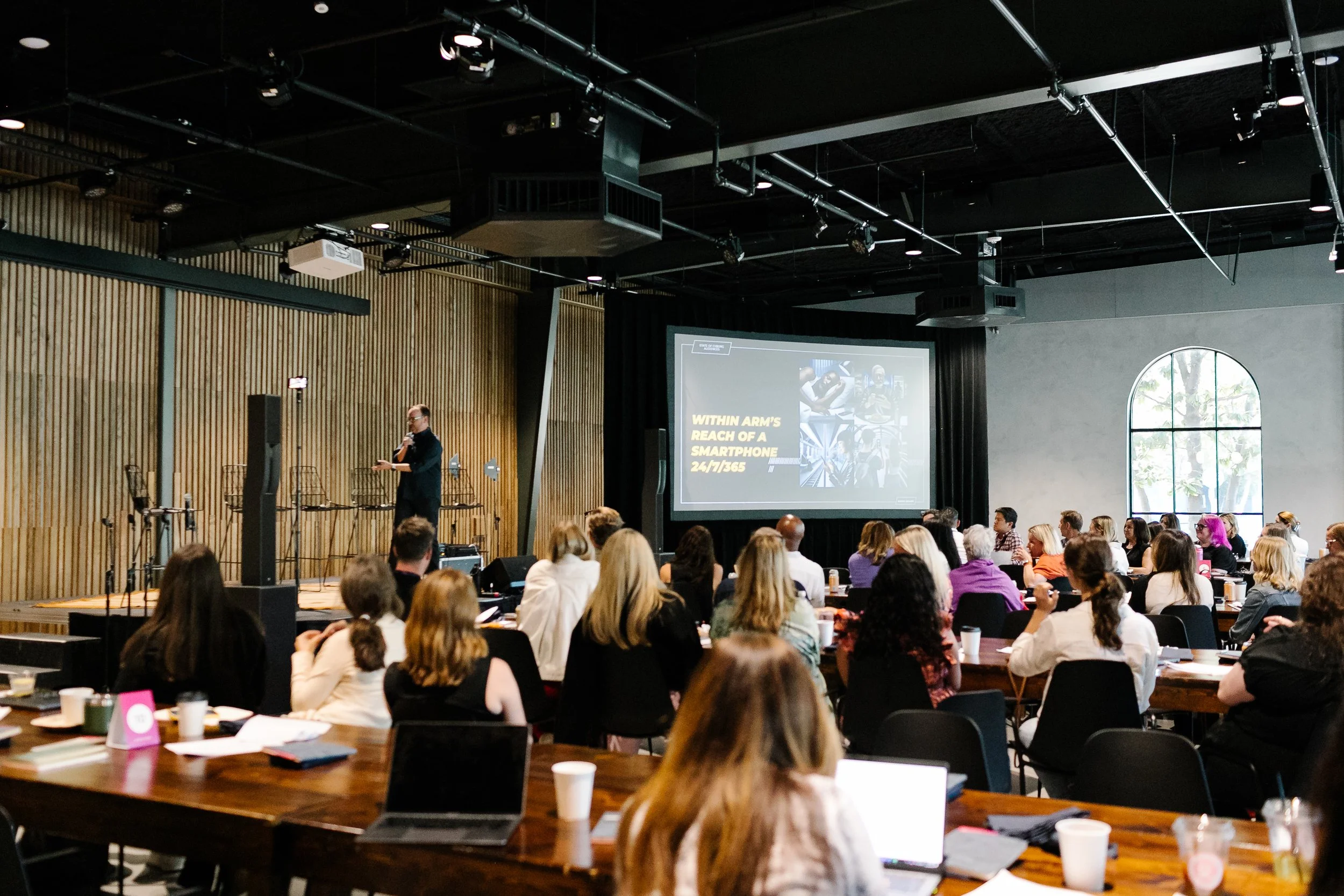 A conference room filled with attendees listening to a speaker on stage, with a large screen displaying a presentation about smartphones.