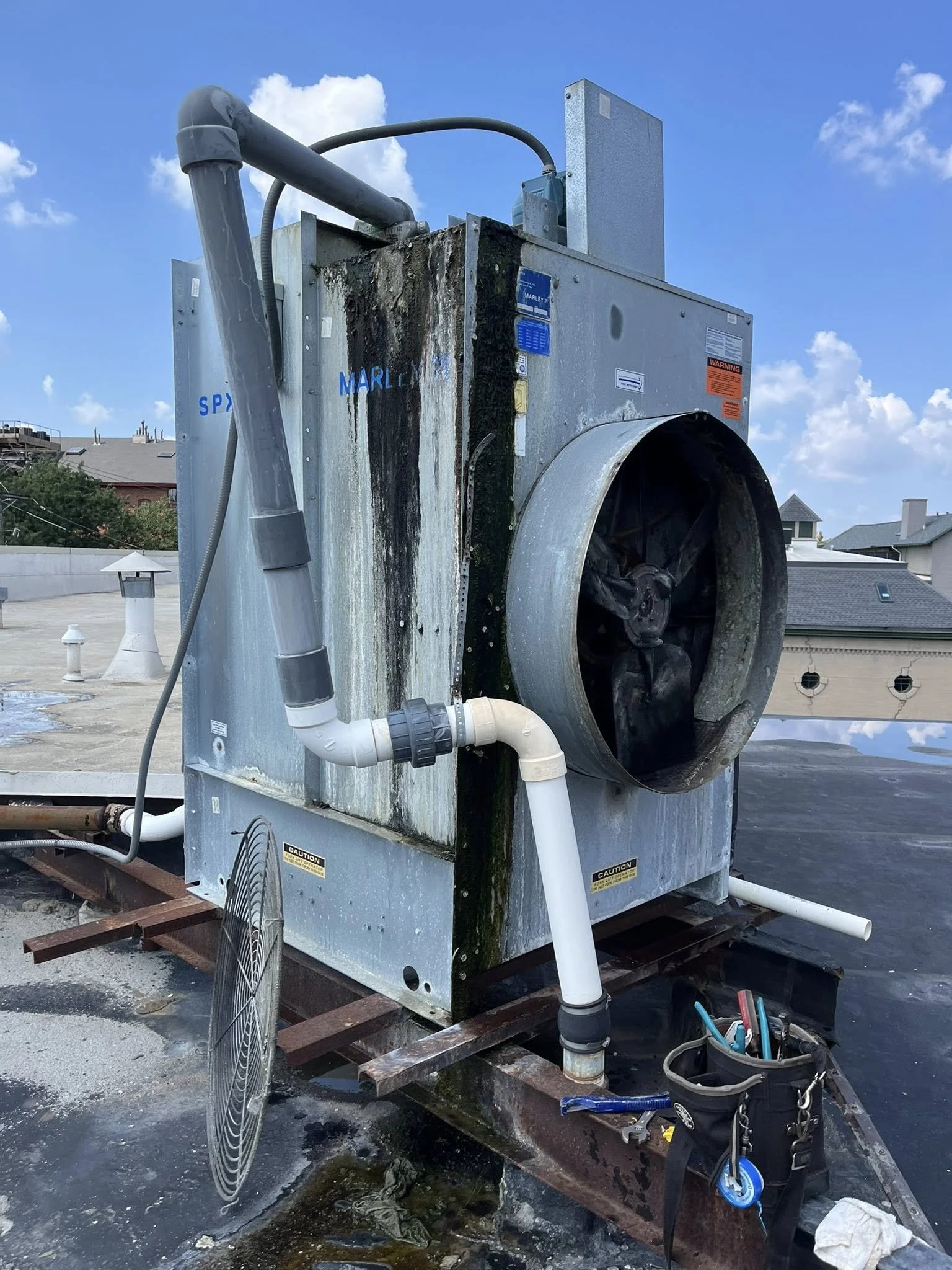 An industrial rooftop air conditioning unit with visible mold and moss, connected to white pipes and a black grille fan, with a utility tool bag and tools nearby, under a partly cloudy sky.