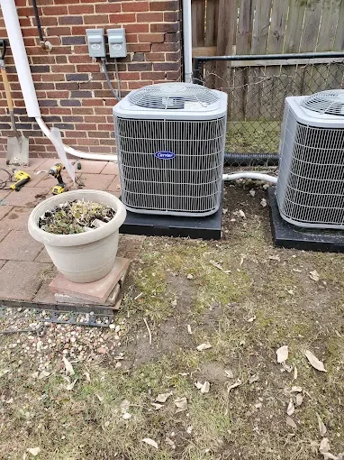 Two outdoor air conditioning units installed on a concrete pad next to a brick building, with a potted plant and some gardening tools nearby.