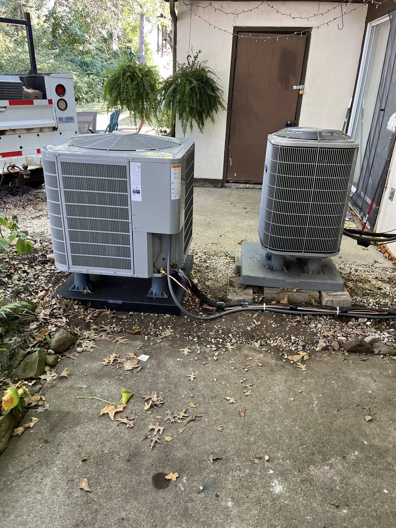 A residential outdoor scene showing two air conditioning units and a trailer with a tree and hanging fern plants in the background.