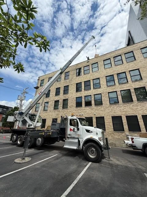 A street scene with a crane truck elevated next to a multi-story brick building, with a person visible at the top of the crane.
