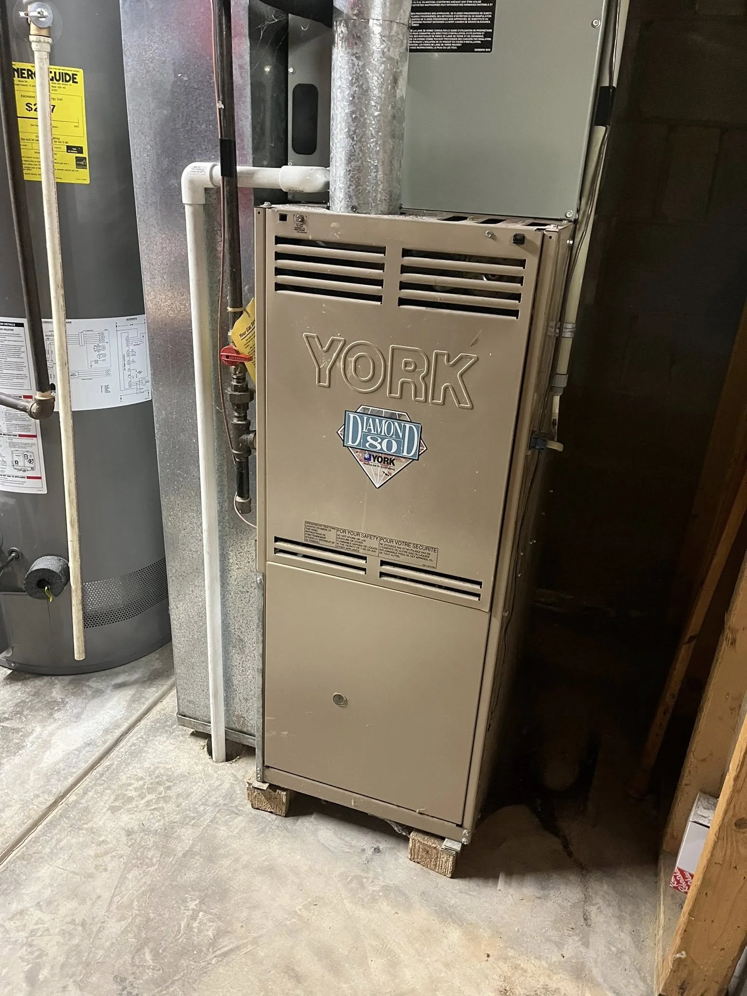 A beige York furnace with a Diamond 80 sticker, located in a basement next to a water heater and to the right of a pipe.