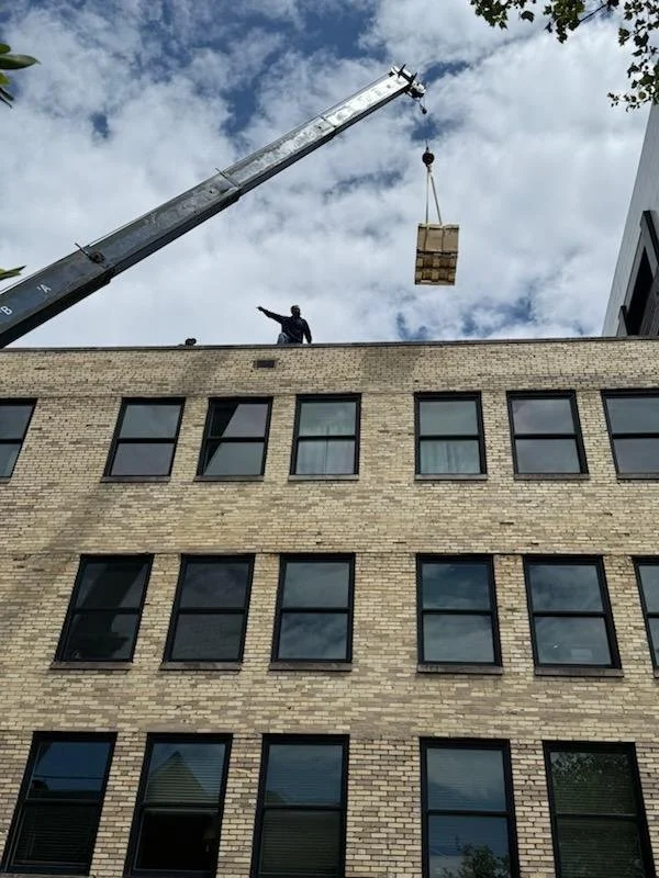 A construction worker standing on the top of a beige brick building, pointing toward a floating pallet suspended in the air by a crane with a blue sky and clouds in the background.