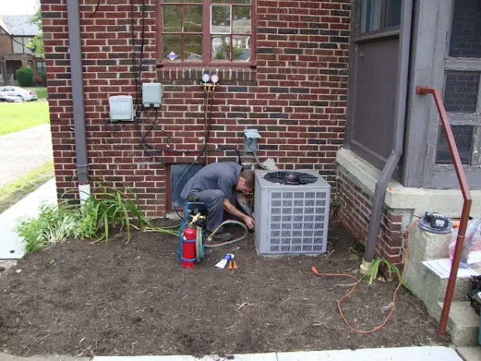 A person working on an outdoor air conditioning unit next to a brick house, with tools and a fire extinguisher nearby.