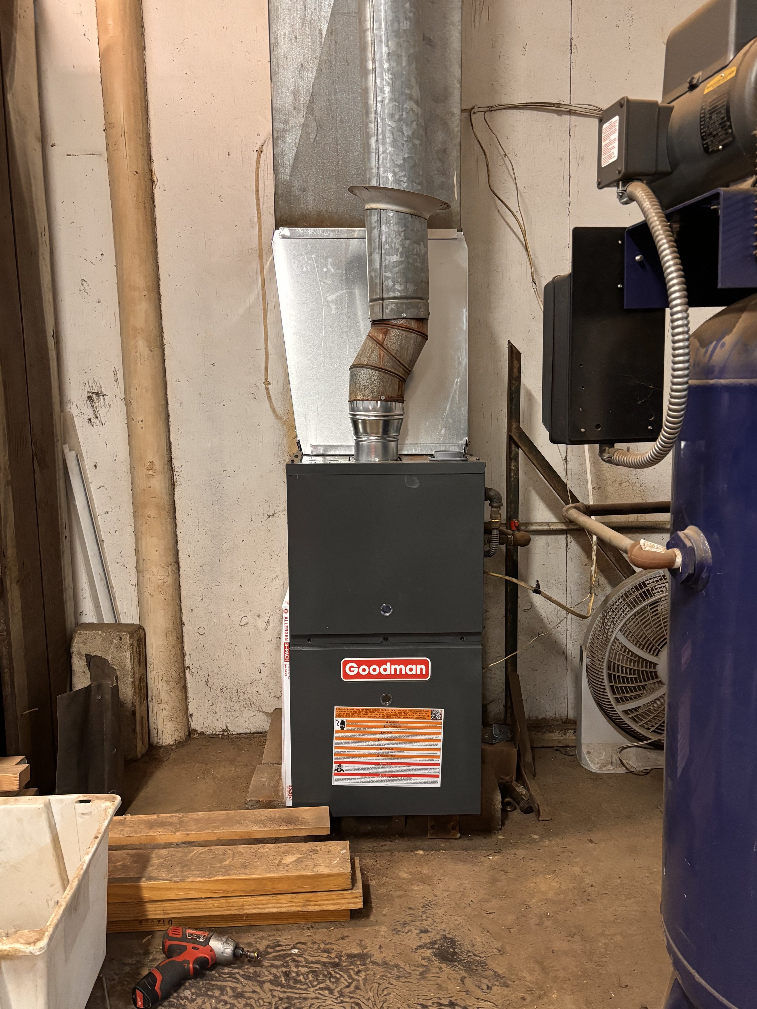 A Goodman furnace in a basement, with a rusted exhaust pipe, surrounded by tools and construction materials.