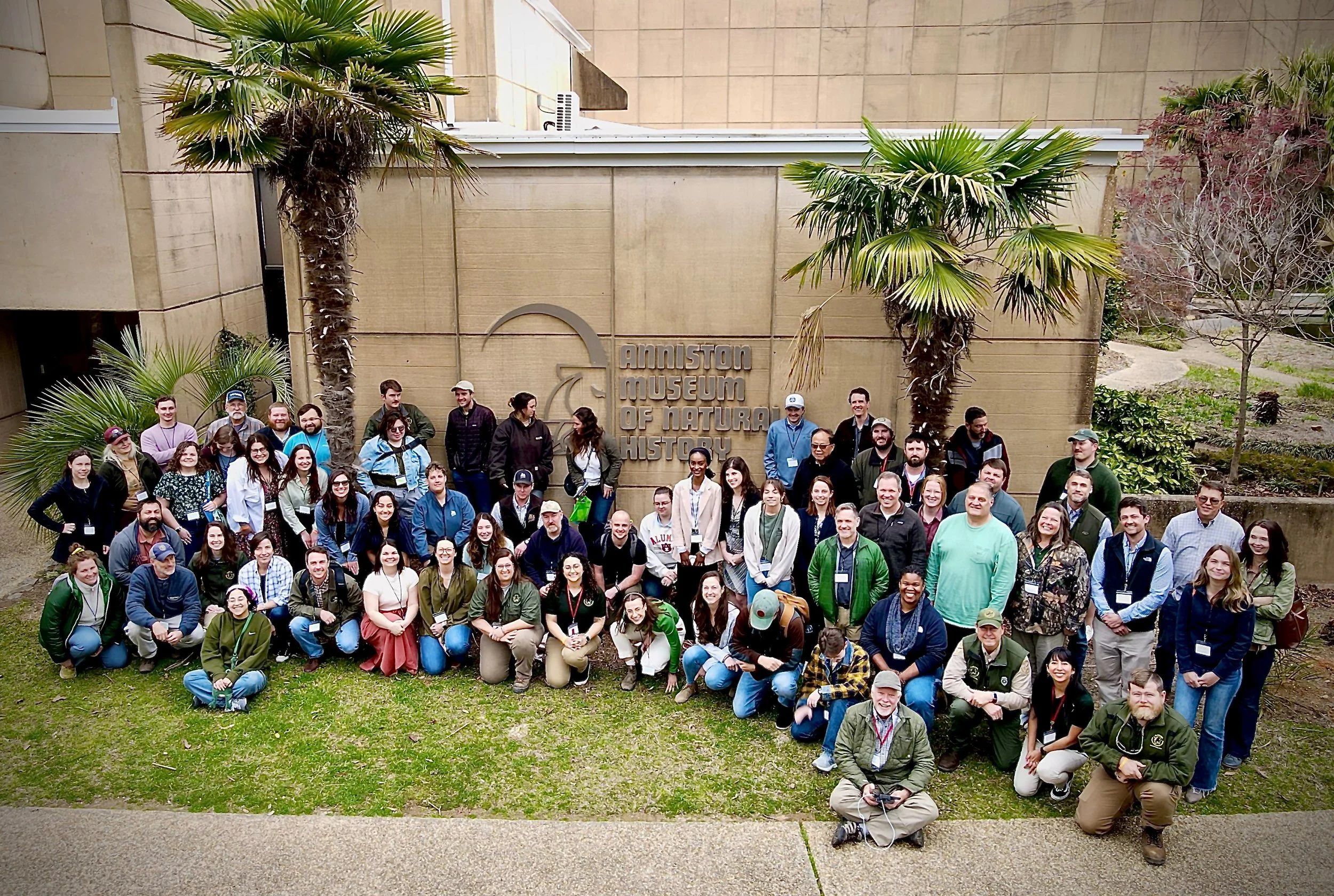 Group of people standing and sitting in front of the Anniston Museum of Natural History, with palm trees and a beige building in the background.