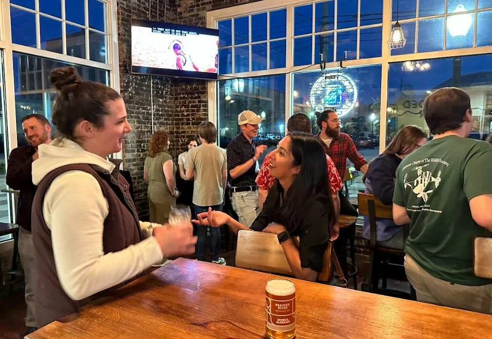 People socializing inside a modern bar or restaurant with large windows, exposed brick walls, and a TV screen. Two women are engaged in conversation, with one holding a drink and laughing.