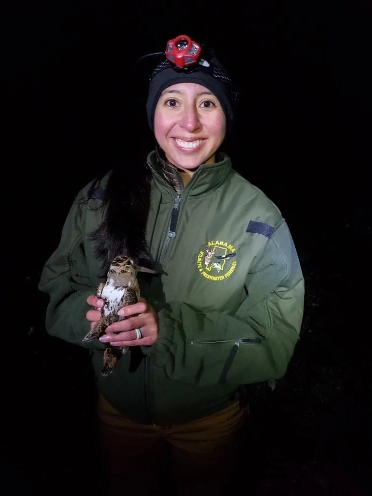 A woman wearing a green jacket with an Alabama logo, a black beanie with a headlamp, smiling and holding a bird, possibly a woodcock, in her hands at night.
