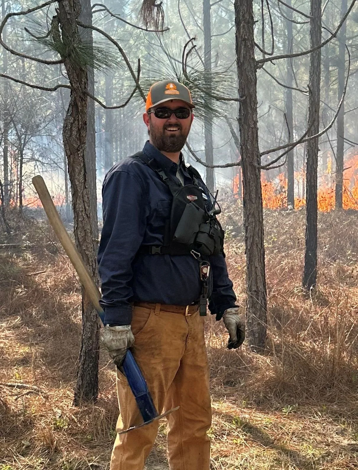 A man standing in a forest fire scene, smiling, holding a shovel, wearing a cap, sunglasses, gloves, and firefighting or forestry gear, with flames visible in the background.
