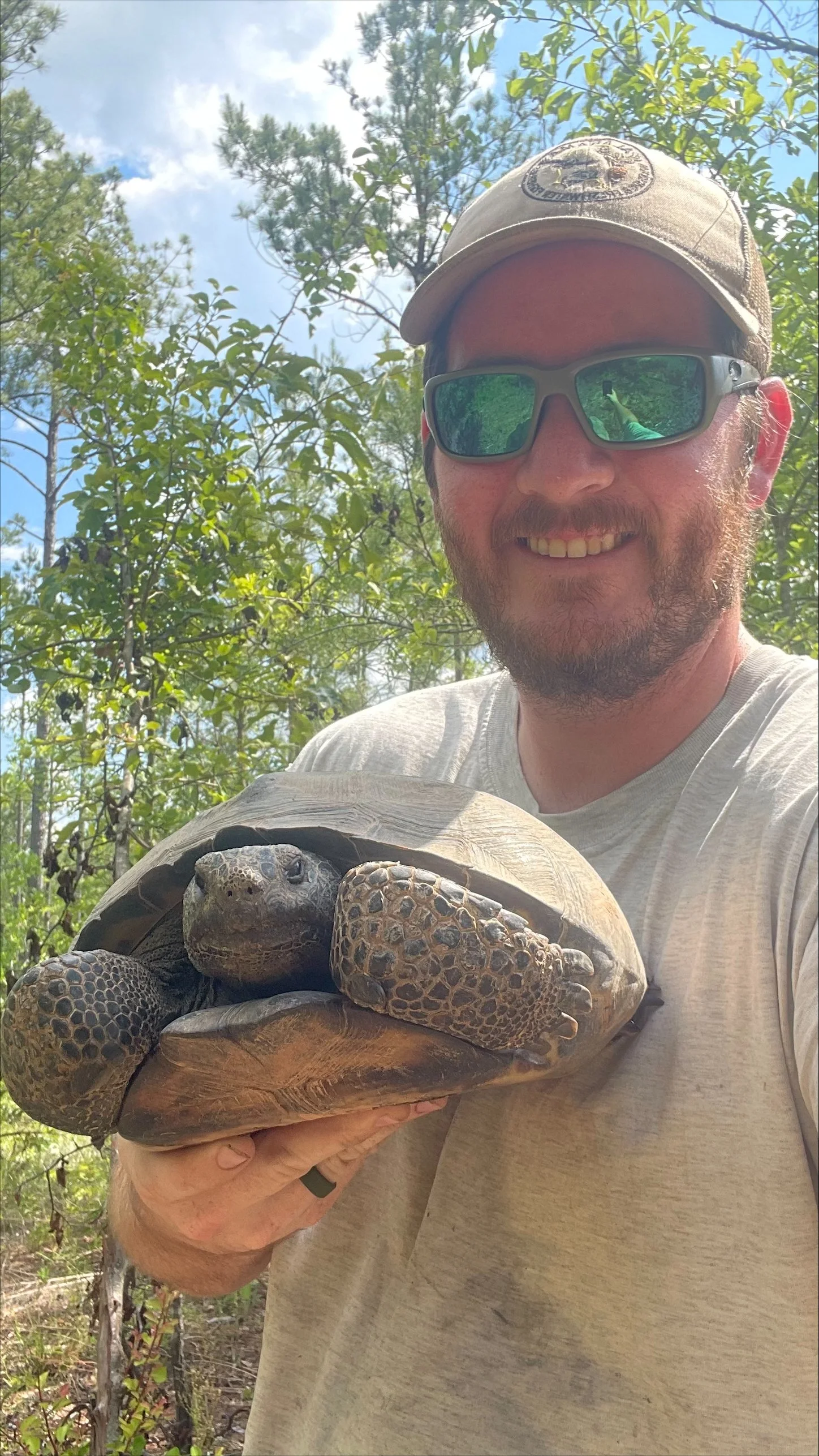 A man wearing sunglasses and a beige cap is holding a large tortoise in an outdoor setting with green trees and blue sky in the background.