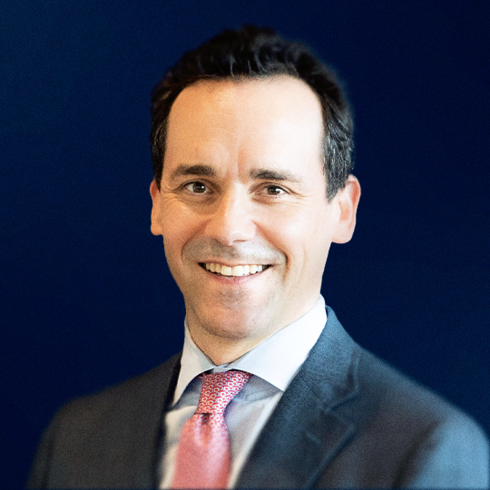 A smiling man in a suit with a pink tie, standing against a dark blue background.