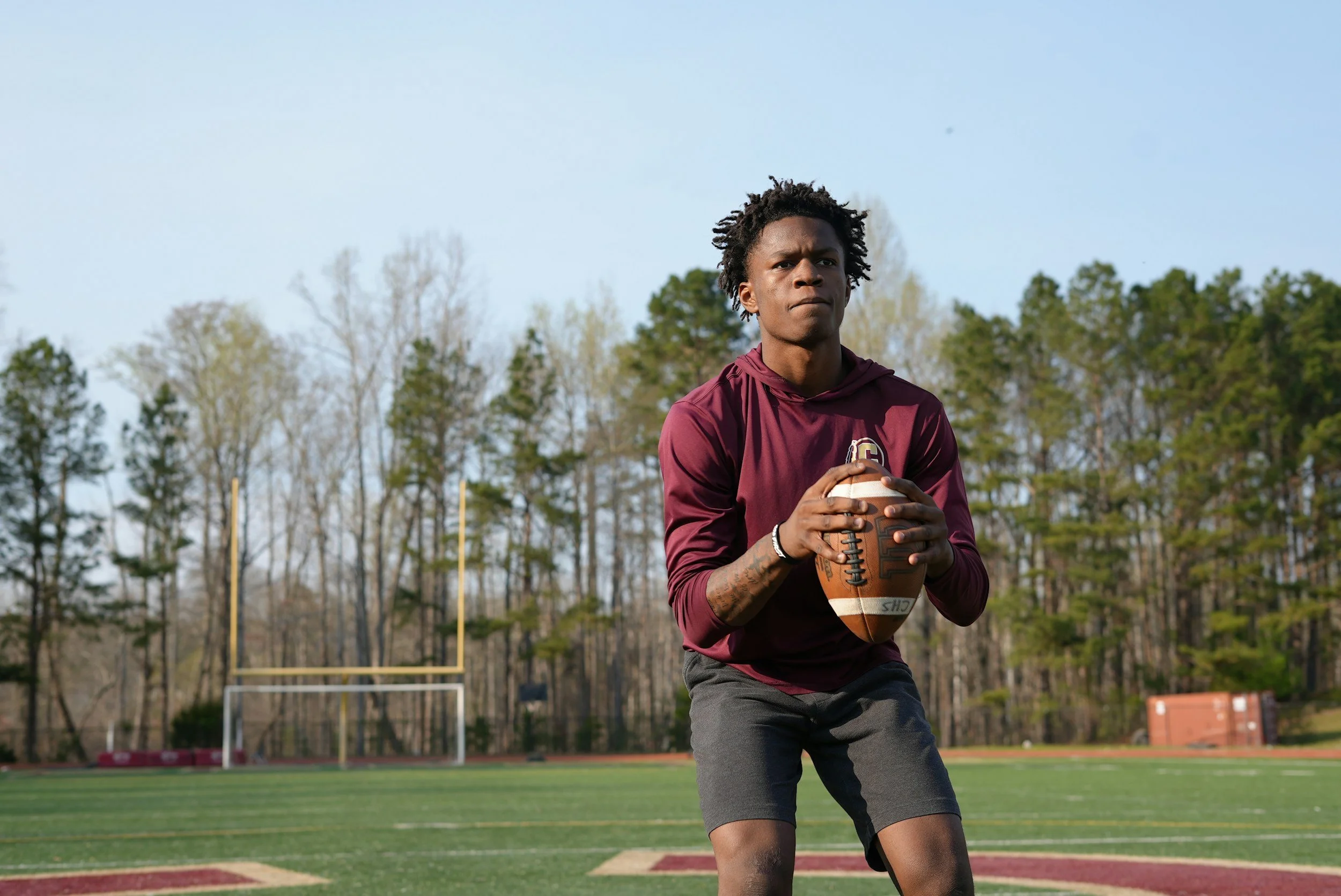 A young man poised to throw a football on a football field with goalposts and trees in the background.