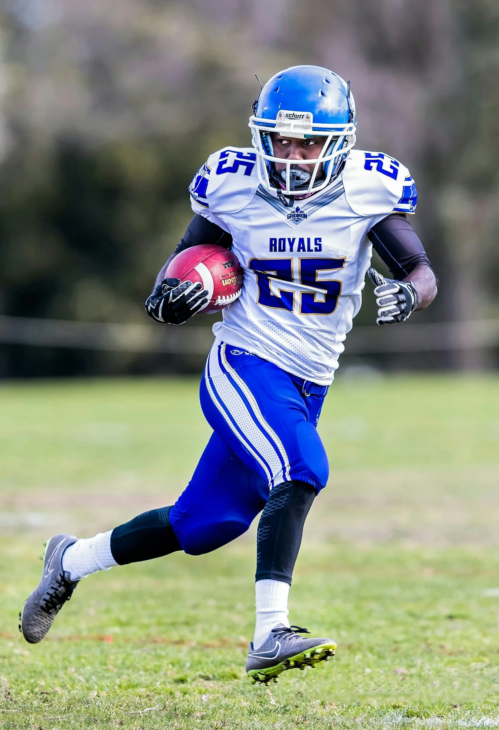 A football player in full gear, wearing a white jersey with blue and gold accents, and the number 25, running with a football during a game.
