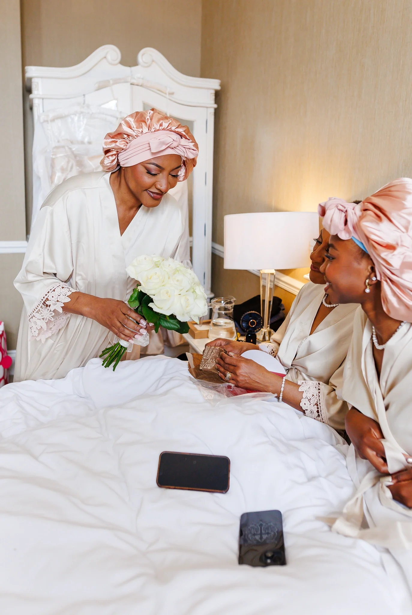 Three women in satin pajamas and matching silk head wraps in a hotel room. One woman in a cream robe presents a bouquet of white roses to another woman sitting on a bed, holding a gift and smiling. The third woman, also in cream satin, looks on, smiling. Smartphones rest on the bed.