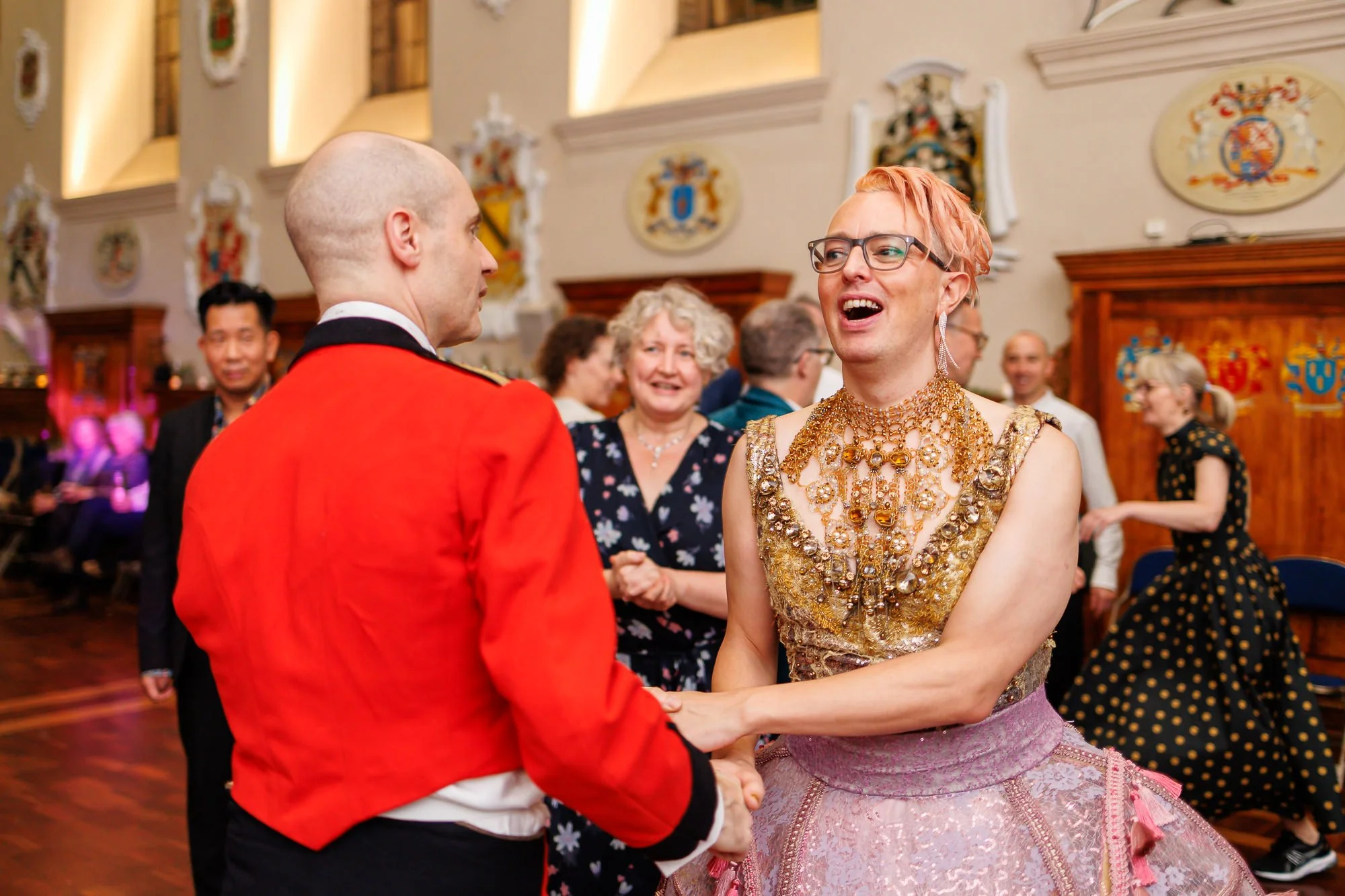 A woman with pink hair, glasses, and elaborate jewelry dances and laughs with a man in a red jacket at a social event in a decorated hall.
