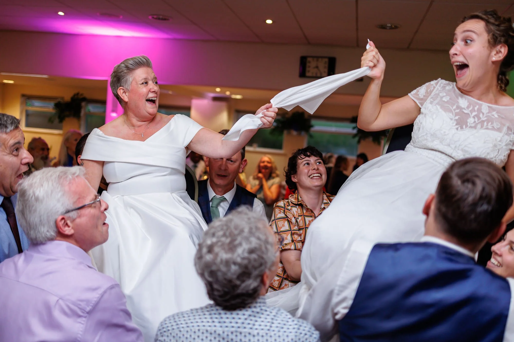 Two brides are holding a hankerchief between them as they are held up on chairs during a hora while everyone celebrates around them in a decorated hall during a lesbian wedding in London.
