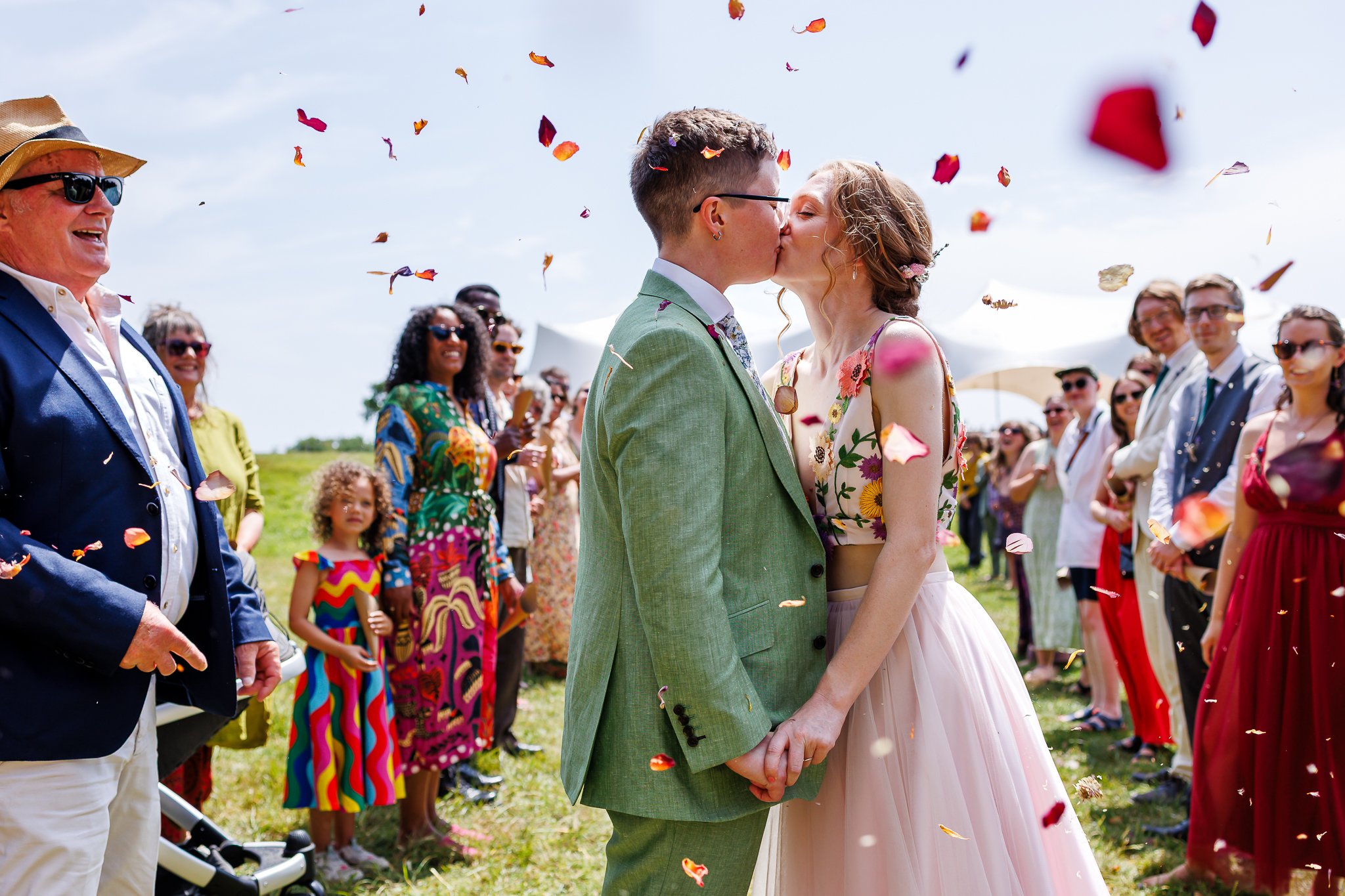Couple in wedding attire kissing, surrounded by friends and family during an outdoor wedding celebration with confetti in the air.