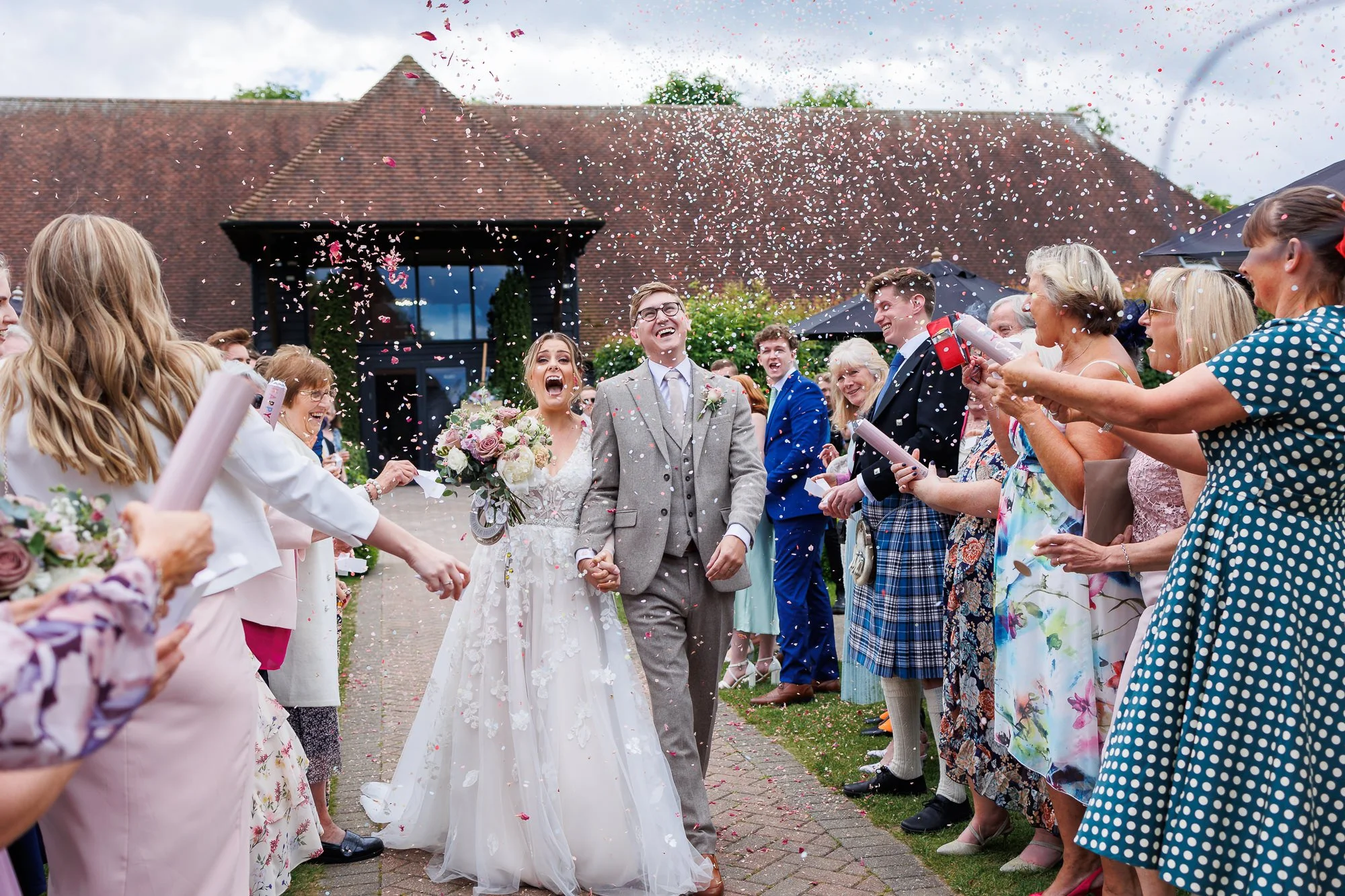 Bride and groom holding hands and smiling as guests throw confetti during their wedding celebration outside a building with a sloped roof.