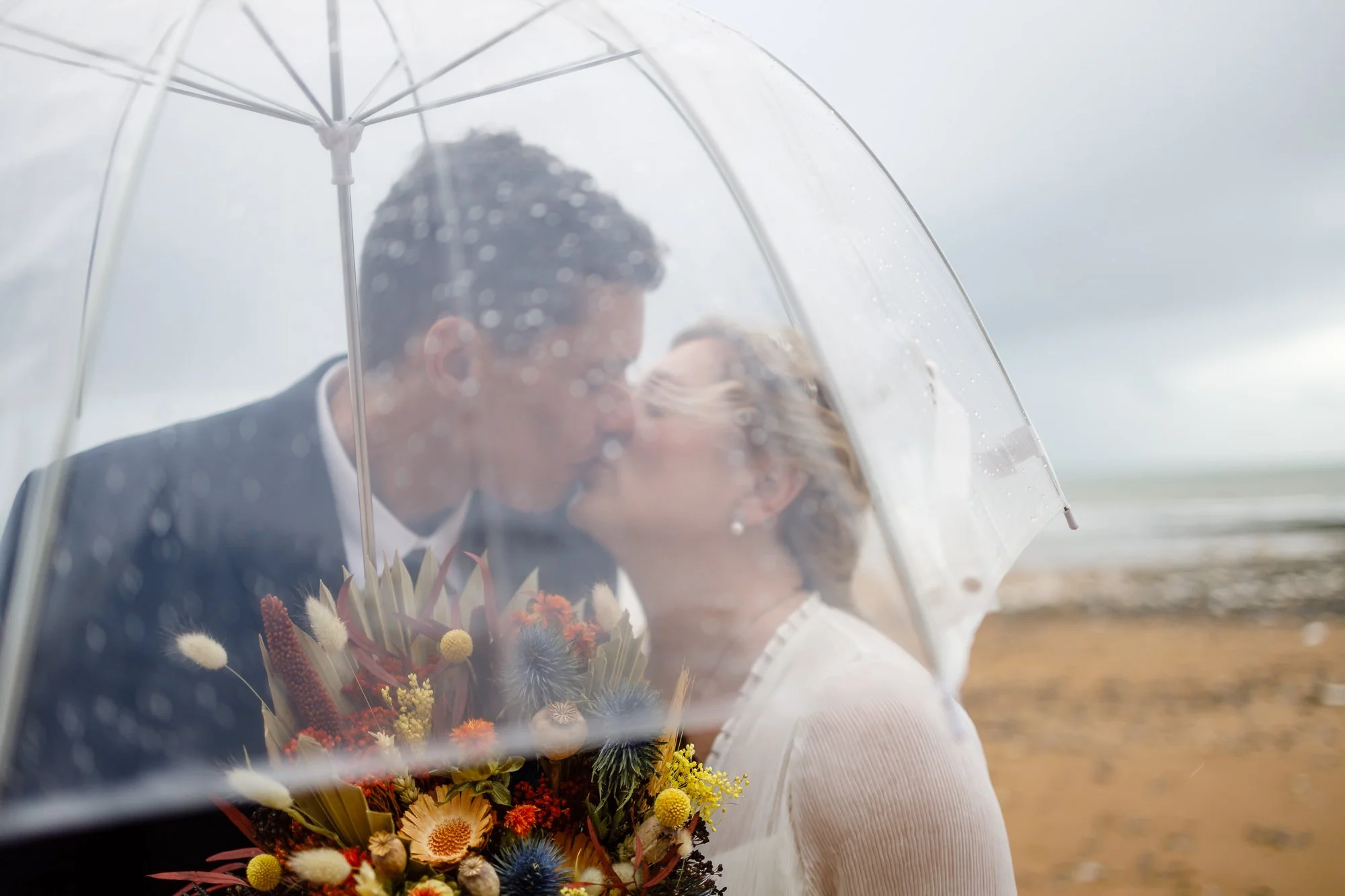A couple kissing under a transparent umbrella on a rainy day at the beach, with the woman holding a bouquet of colorful flowers.