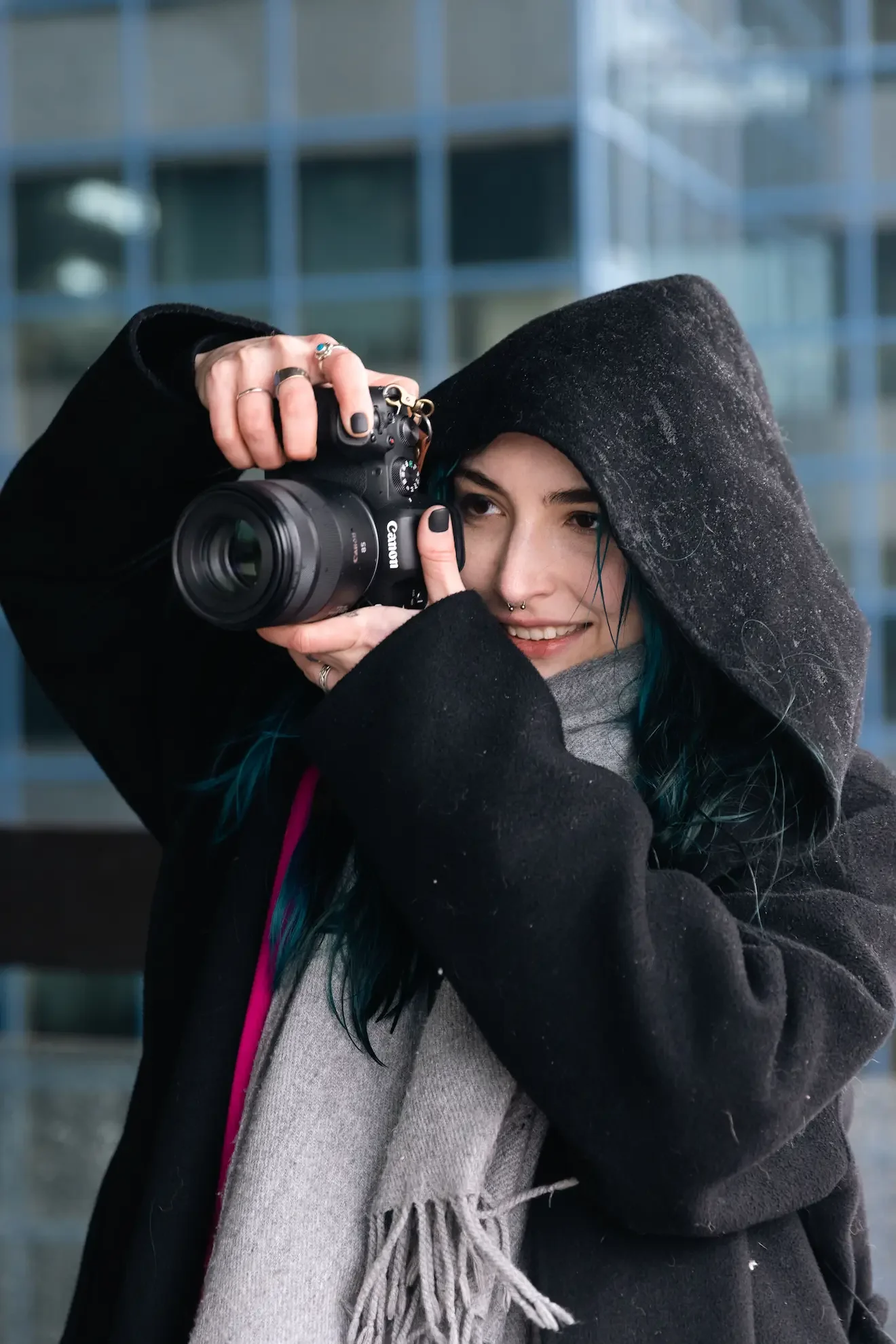 A young woman in a black hoodie and grey scarf takes a photograph with a Canon camera outside an office building.