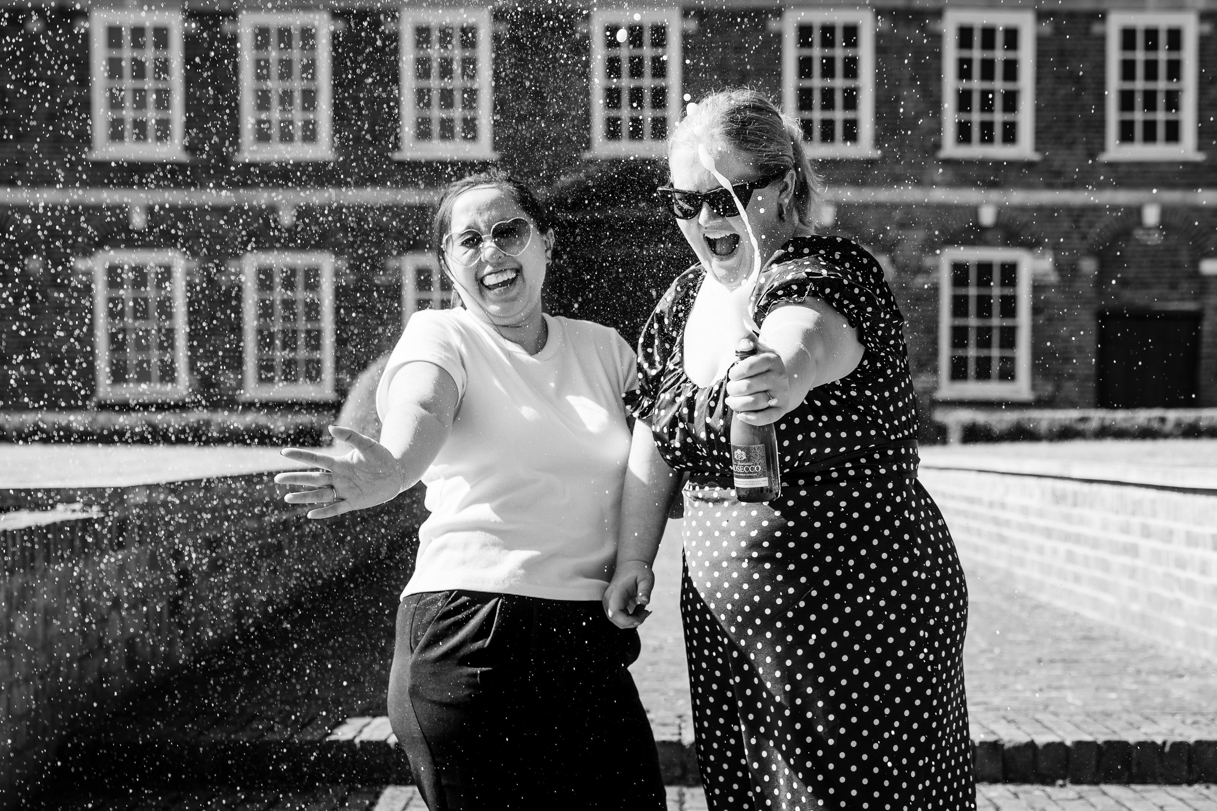 Black and White photo of queer couple laughing and spraying champagne from a bottle at Hall's Place, London.