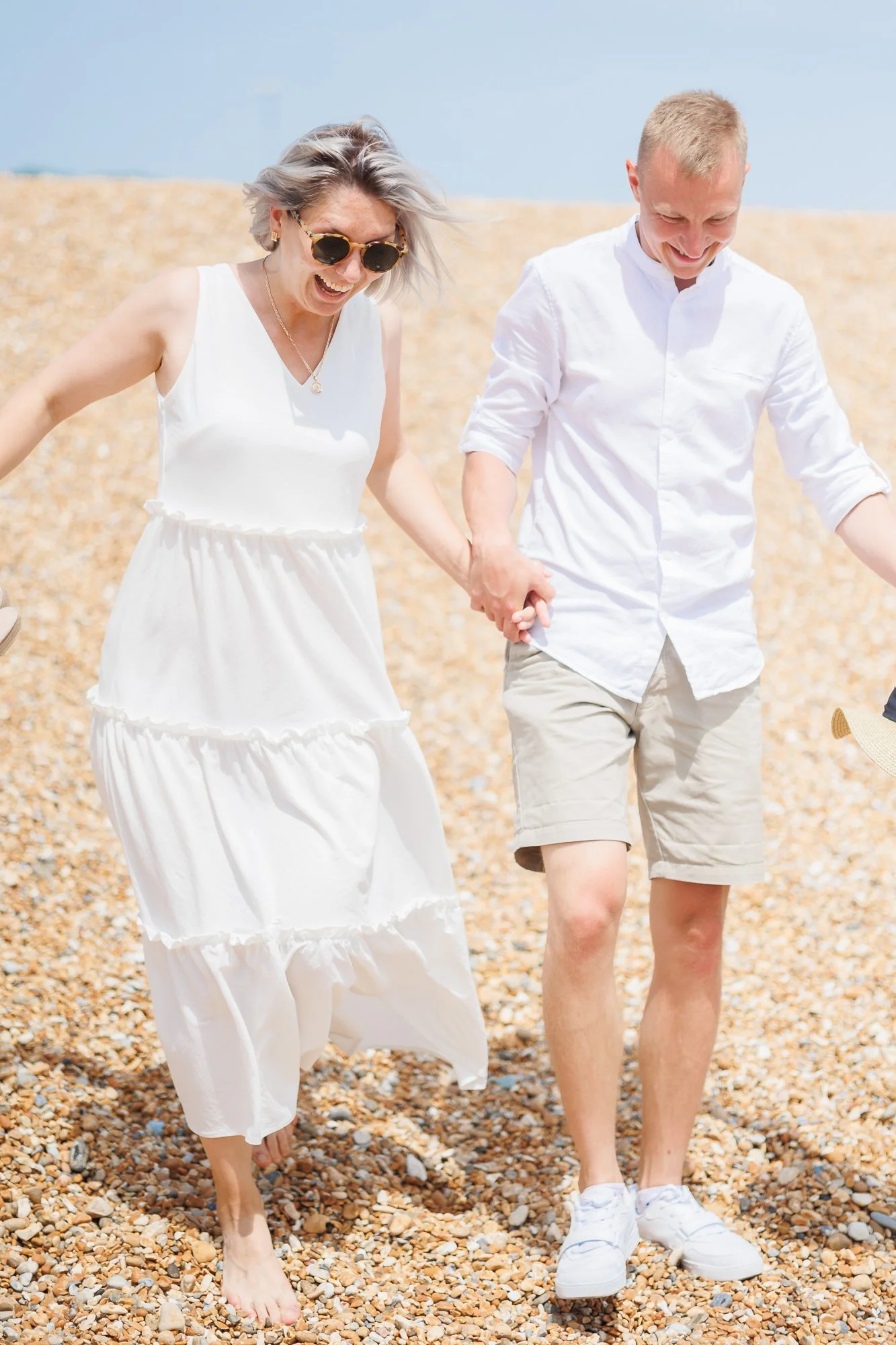 A woman and a man holding hands and walking on a pebble beach, smiling and enjoying a sunny day.