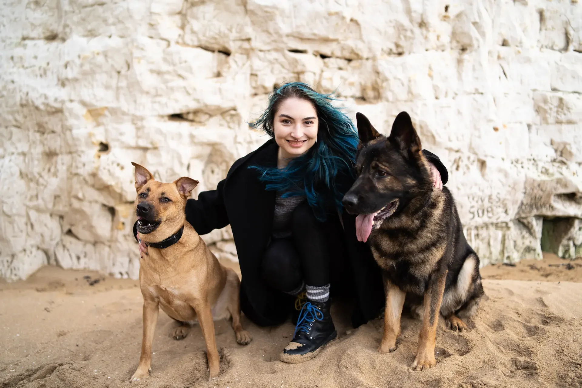 A young woman with blue streaks in her hair crouching on sandy ground between two dogs, one tan and one black with brown markings, in front of a beige rock wall.
