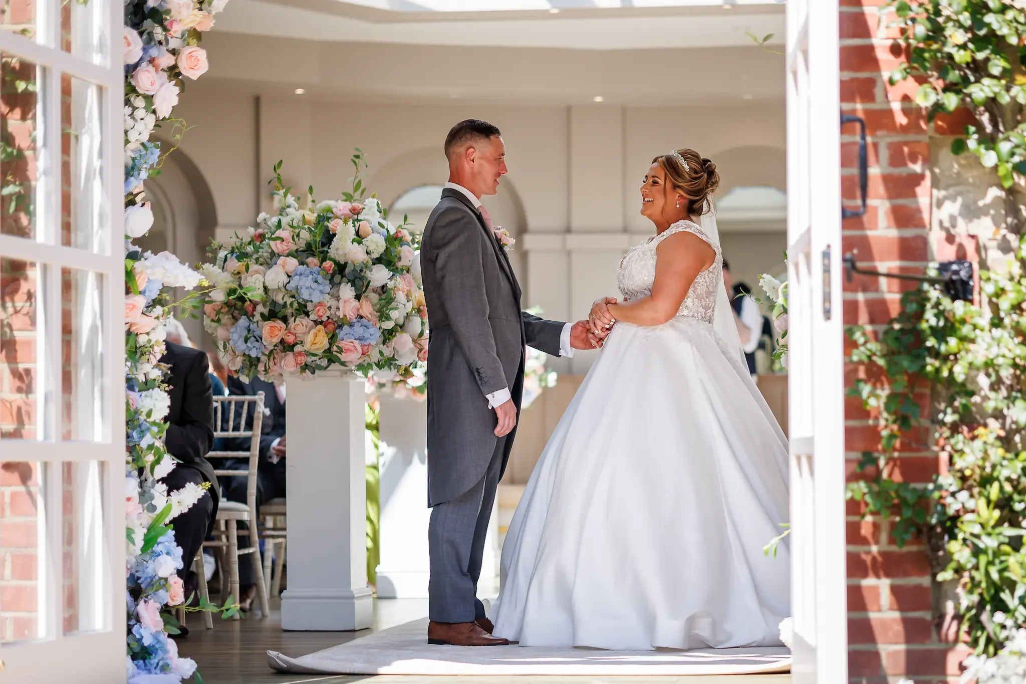 Couple during ceremony in The Garden Room at The Orangery in Maidstone. The view is from the outside through the doors that are open, the couple are looking at each other and smiling,