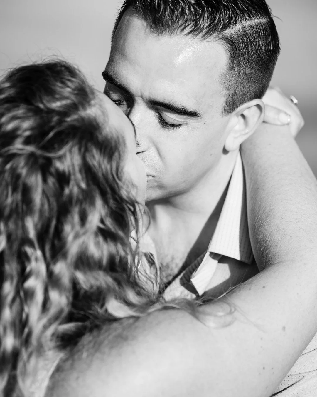 Black and white photo of a man with dark short hair kissing a woman whose arms are around his neck. The woman's hair is curly. This is a proposal photoshoot on the White Cliffs of Dover.