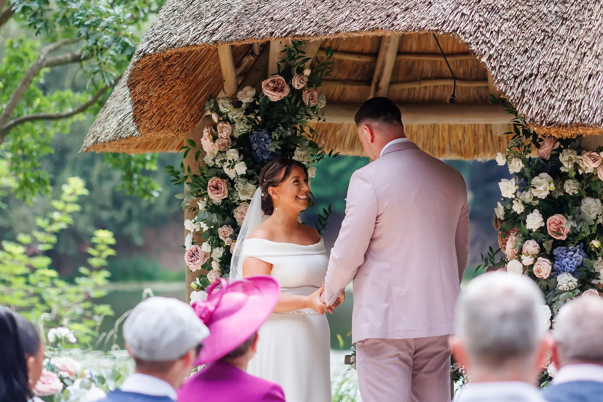 Couple smiling at each other during a wedding ceremony at The Lapa at The Orangery in Maidstone which is a thatch roof structure with flower arches.