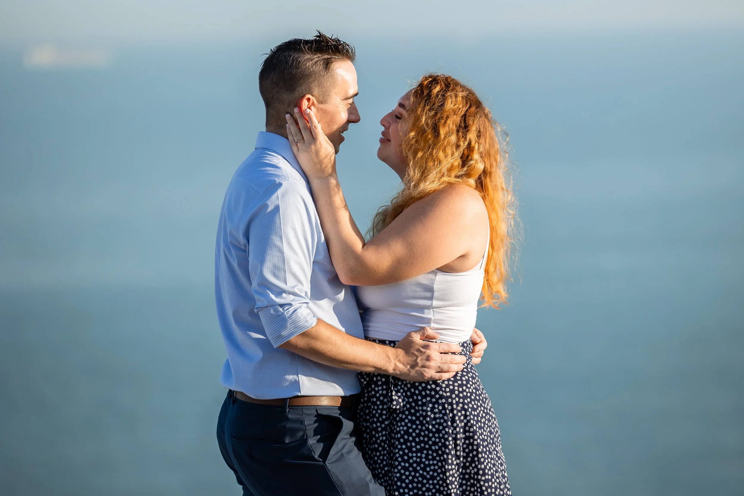 A man and a woman are standing facing each other with the man's hands on the woman's hips and she is holding his face. You can see an engagement ring on her finger. There is a ferry far in the background as this is a proposal on the White Cliffs.