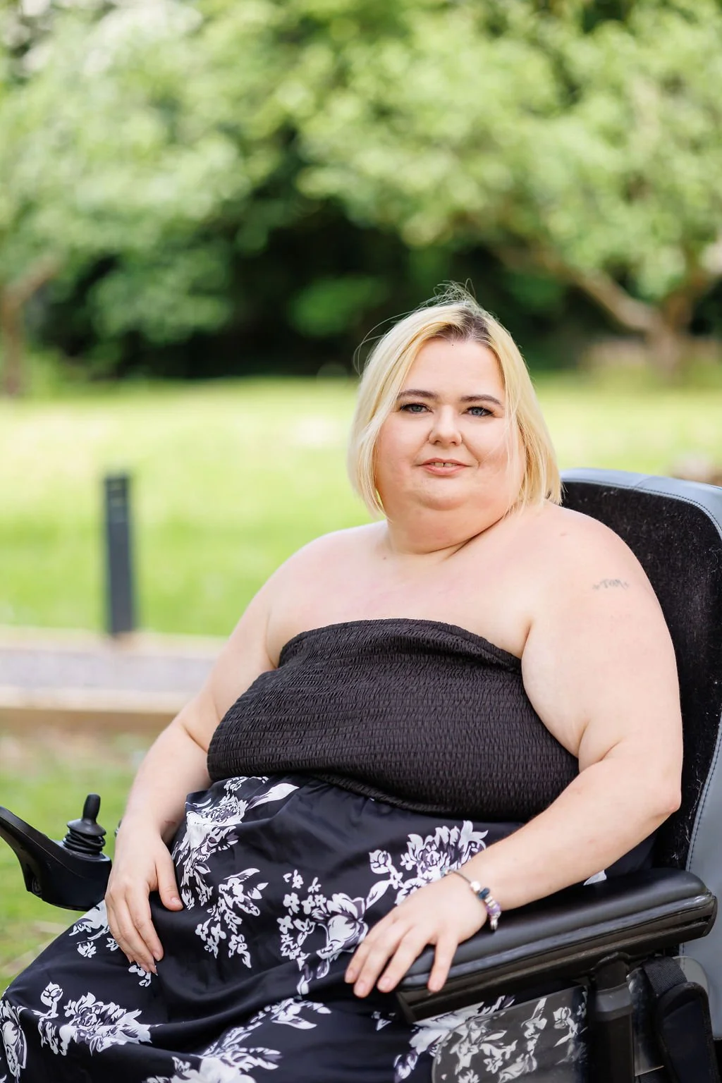 A woman with blonde hair sitting outdoors in a wheelchair, wearing a black strapless top and a floral skirt, with a background of green trees and grass at the Lake House wedding venue in Kent where Nastia Photography is a recommended supplier.