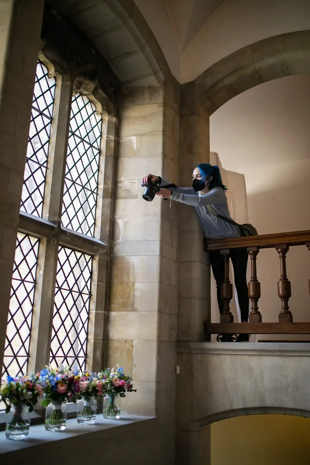 A woman with blue hair wearing a black face mask, gray jacket, and carrying a backpack, stands on a balcony in a historic building, taking a photograph through a lattice-window with a DSLR camera. There are four vases with colorful flowers on the windowsill.
