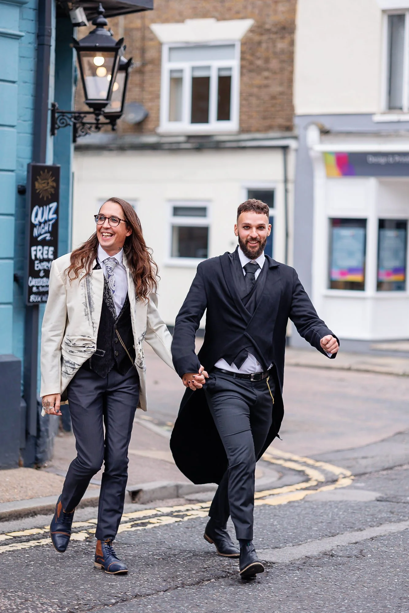 Two people, a woman with long wavy hair and a man with a beard, wearing formal attire, holding hands and running on a city street at dusk, smiling and laughing.