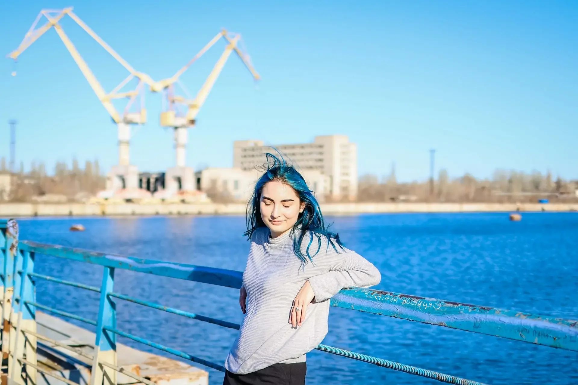 Young woman with blue hair standing by a railing near a body of water, with a cityscape and industrial structures in the background on a clear, sunny day.