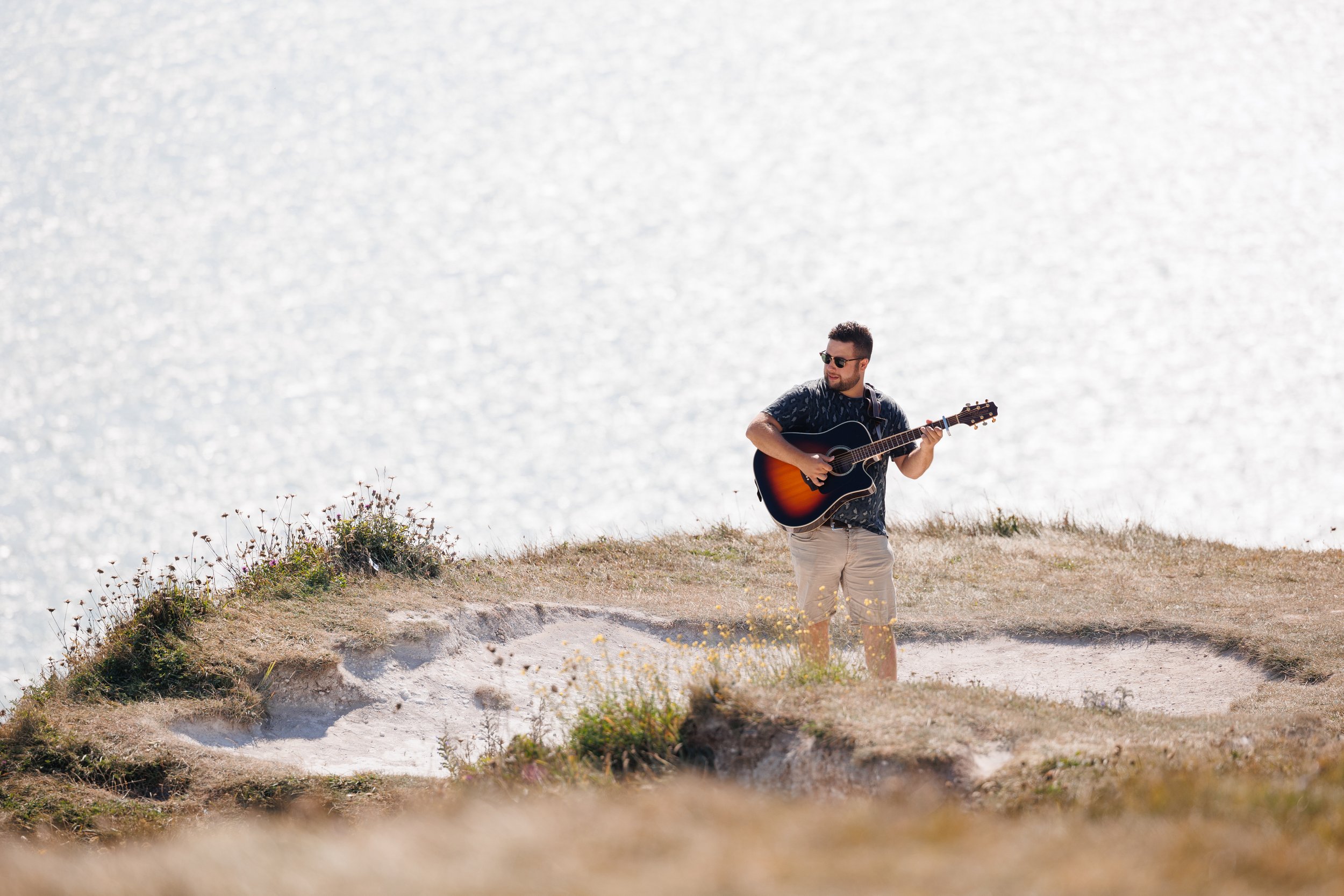 A man wearing sunglasses, a dark patterned shirt, and beige shorts, playing an acoustic guitar on a cliff near water, with sunlight reflecting off the water in the background. This is a musician's branding photoshoot at the White Cliffs of Dover.