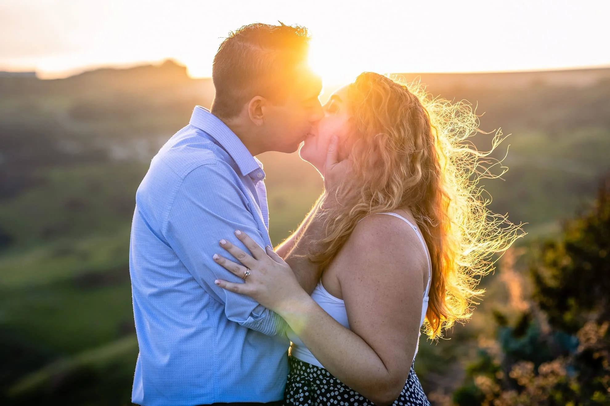 A sunset photo of a man kissing a woman with the sun coming in between them. Her curly hair seems golden and is moving in the wind. You can see an engagement ring on her arm resting on his arm. There are buildings and fields in the background.