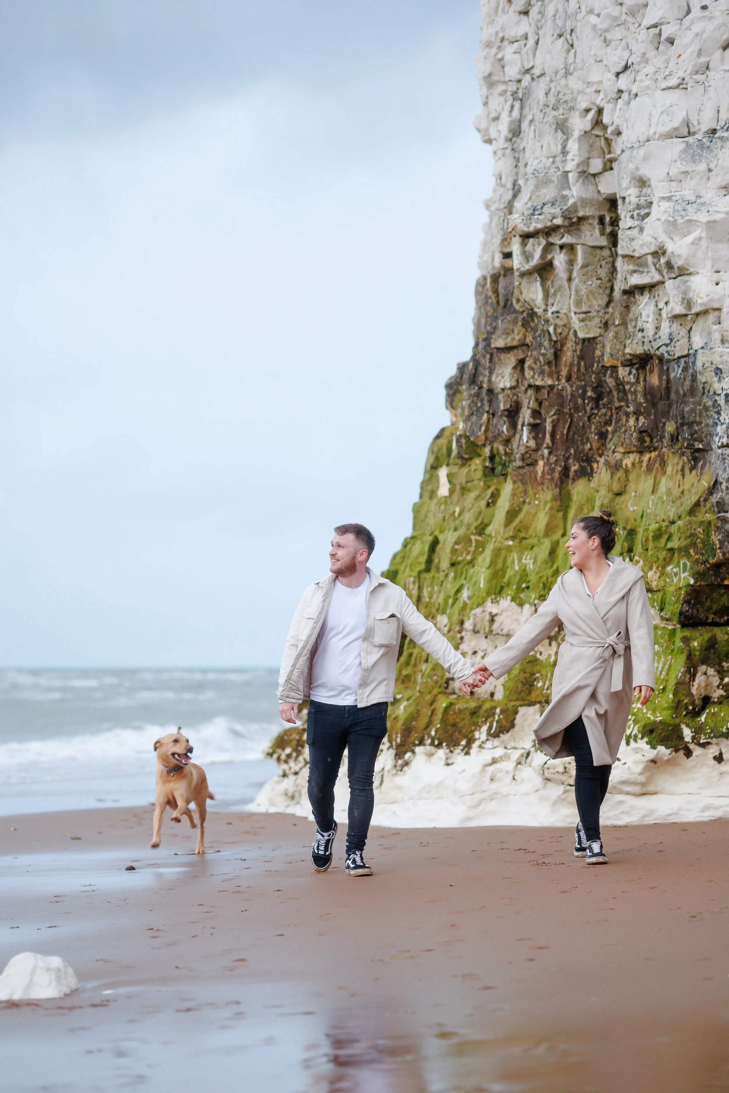 A couple walking hand in hand along the beach with their dog during daytime. The man has short hair and a beard, wearing a light-colored jacket and jeans. The woman has dark hair tied up, dressed in a long beige coat. A sandy beach with a large cliff covered in moss and graffiti, and the ocean in the background.