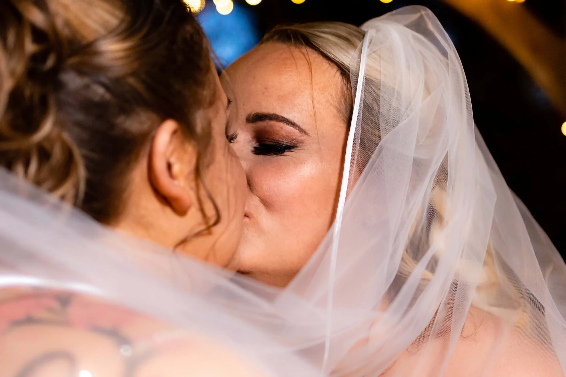 Close-up of two women kissing, one wearing a wedding veil, at a wedding or special event.