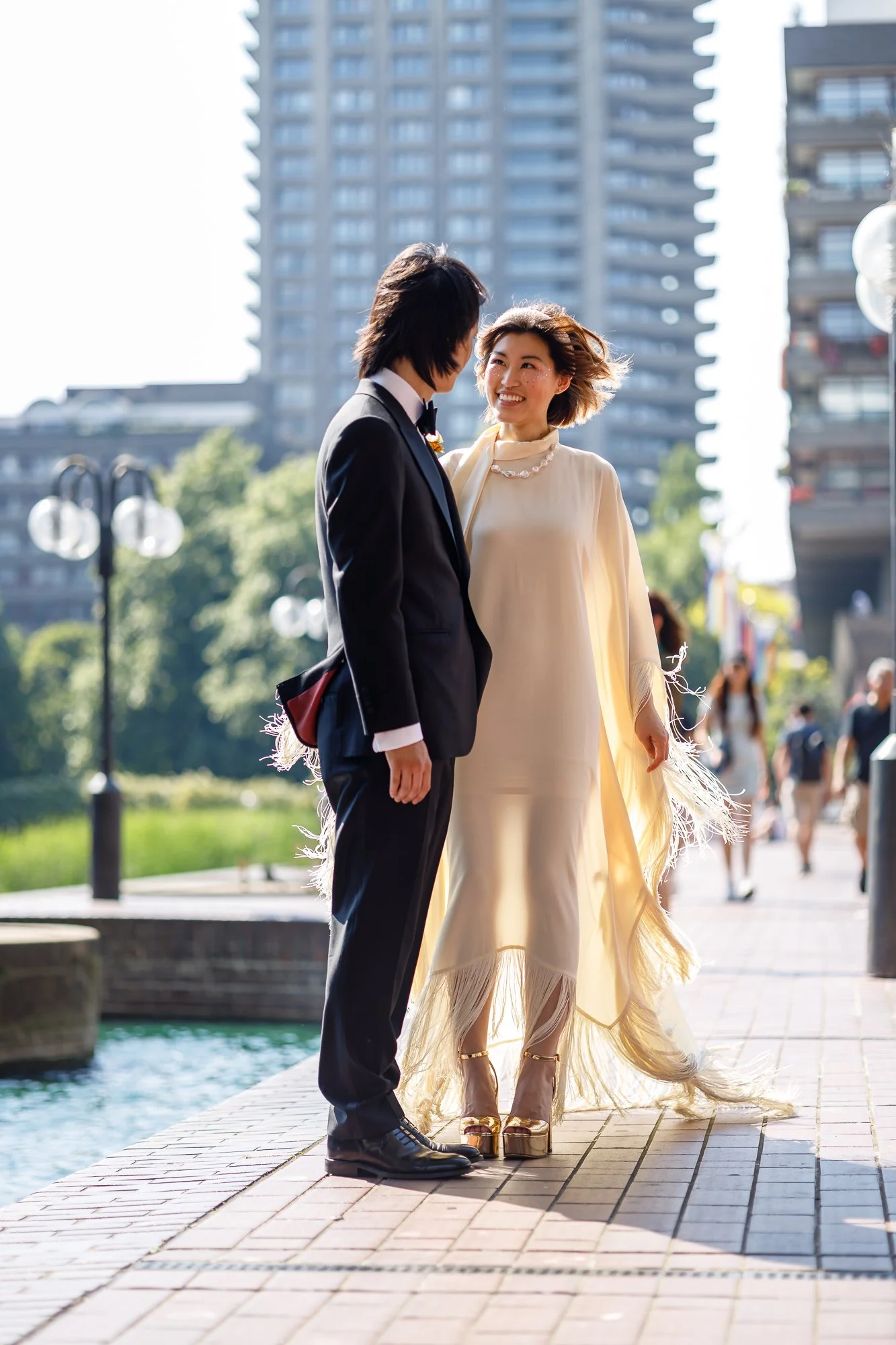 A woman and a man dressed in formal attire standing outdoors in an urban area, smiling at each other with modern high-rise buildings in the background.