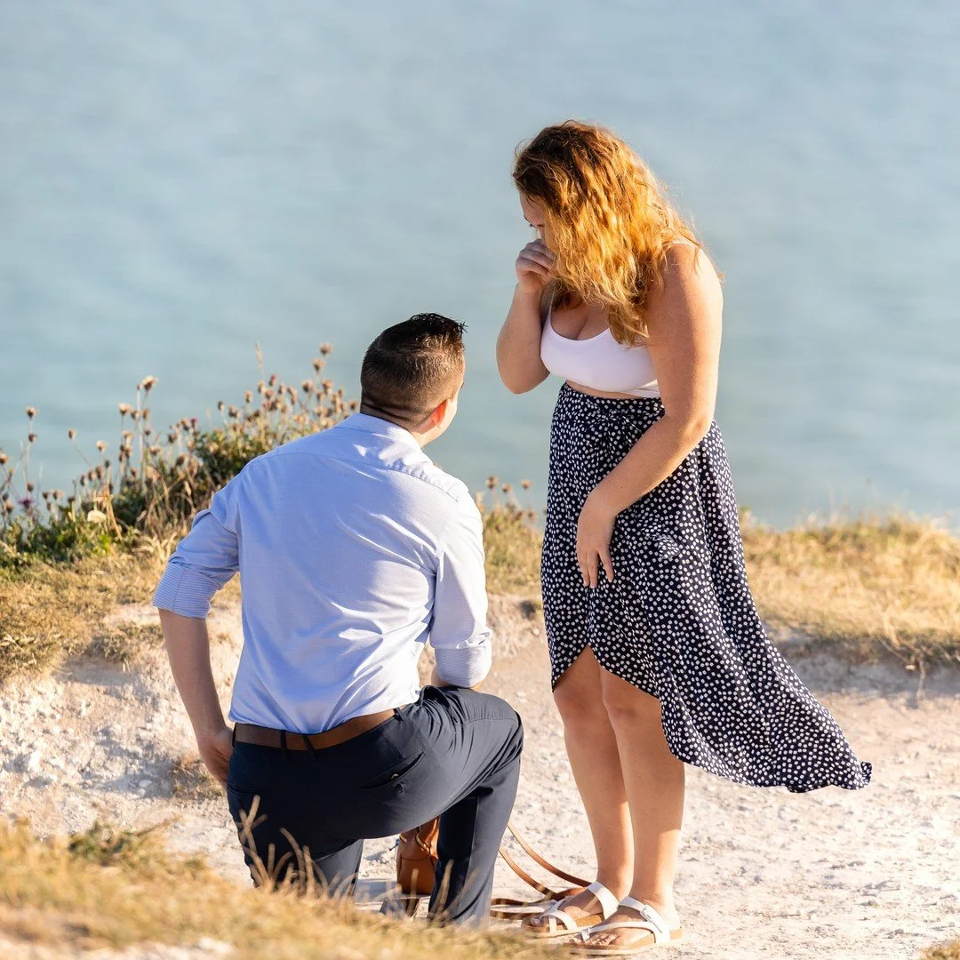 A man in smart dark trousers and light shirt is on one knee proposing at the White Cliffs of Dover to a woman with blonde hair wearing a white tank top and black and white polka dot skirt. The sea is in the background.