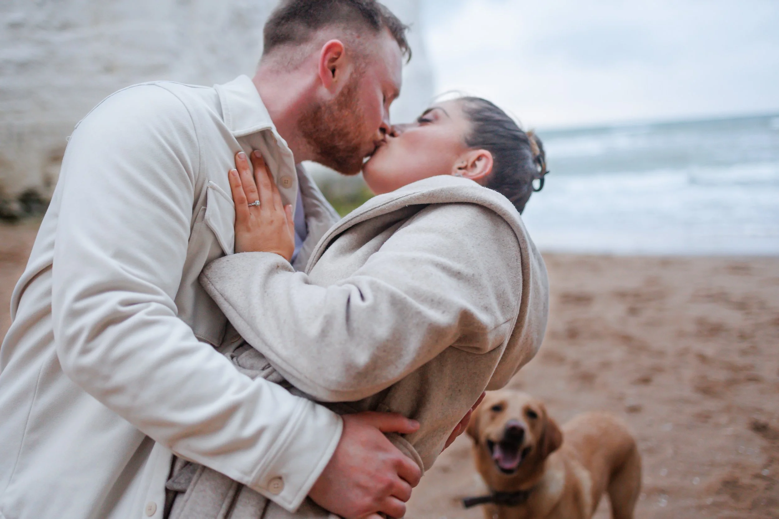 A man and woman are kissing on a beach. The woman is wearing a beige coat, and the man is wearing a white jacket. A dog is in the background, looking at the camera with its mouth open.