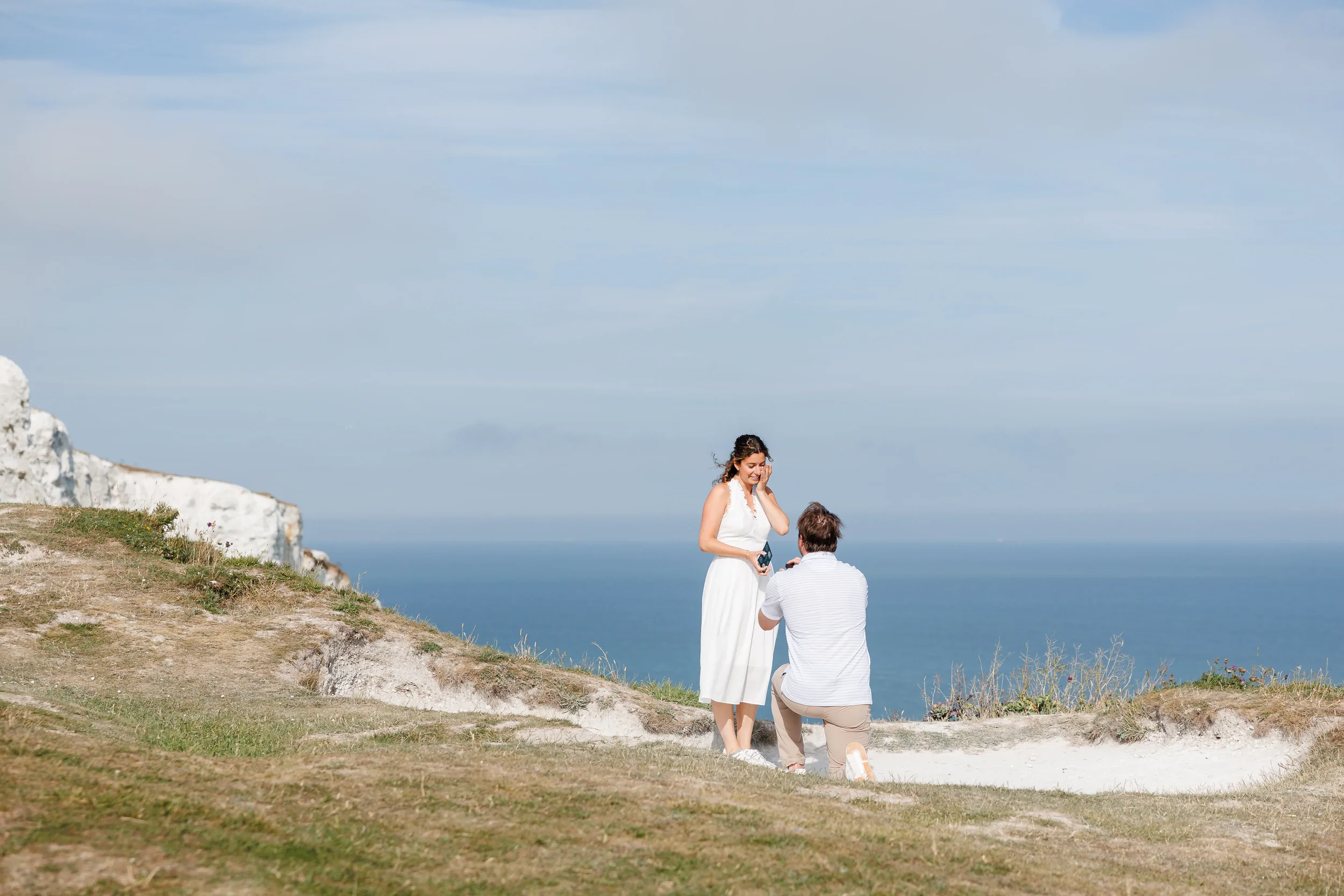A man in light polo shirt and beige trousers proposing on the White Cliffs of Dover to a woman in a white midi dress
