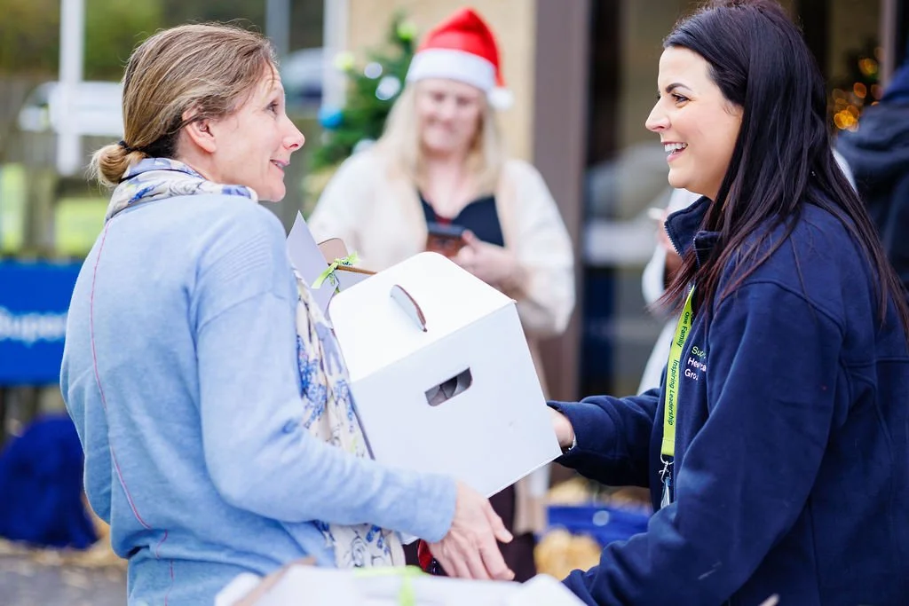 Two women exchanging a white box at a Christmas outdoor event by Superior Healthcare, with a woman wearing a Santa hat in the background.