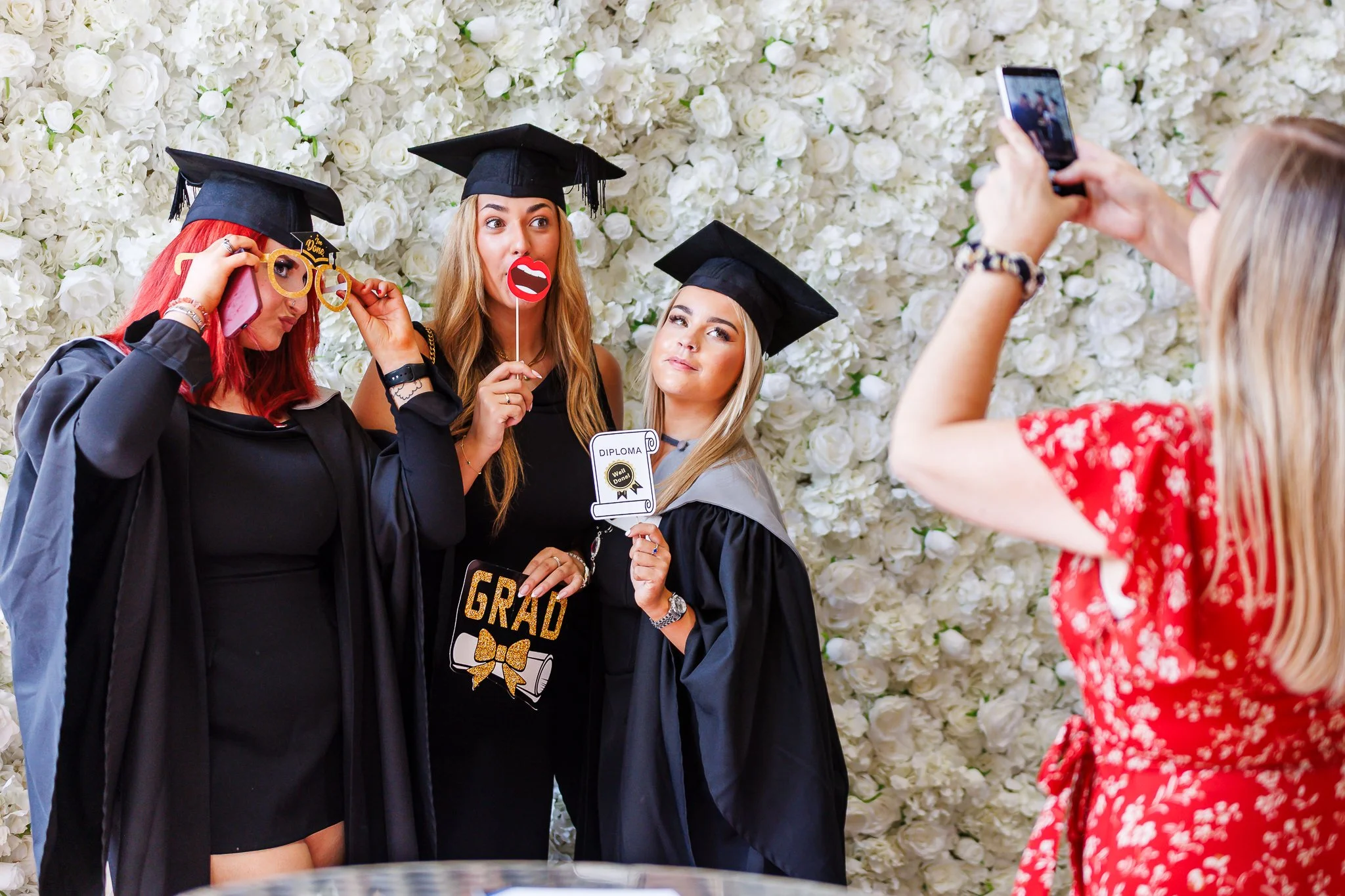Three women in graduation caps and gowns celebrating their graduation from the university of Kent, with one holding a diploma and a sign that says "Grad" in front of a white flower wall, while a person takes their photo.