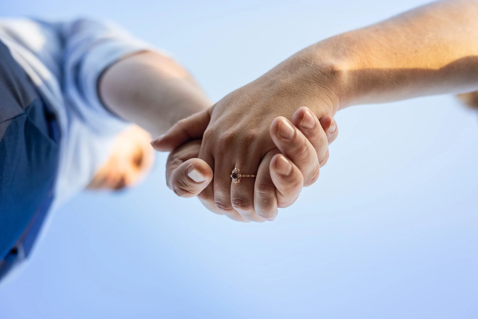 A photograph of hands held from the ground up. You can see a man's silhouette. The focus is on the two hands, one is wearing a gold engagement ring with diamonds and a dark stone in the middle. The background is the blue sky.