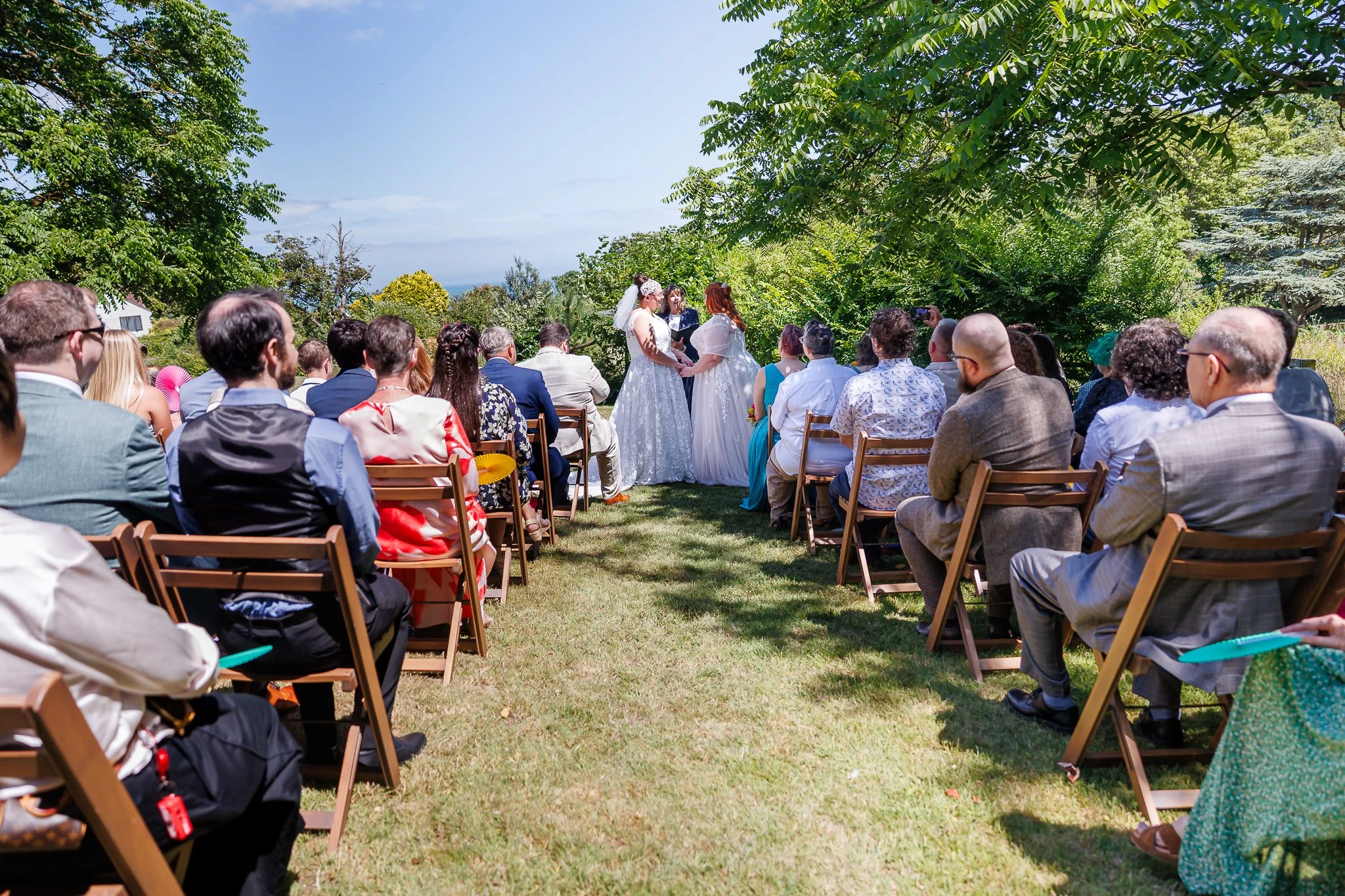 Two Brides in an outdoor ceremony area with guests seated on either side at Pines Calyx in St Margaret's with sea in the background